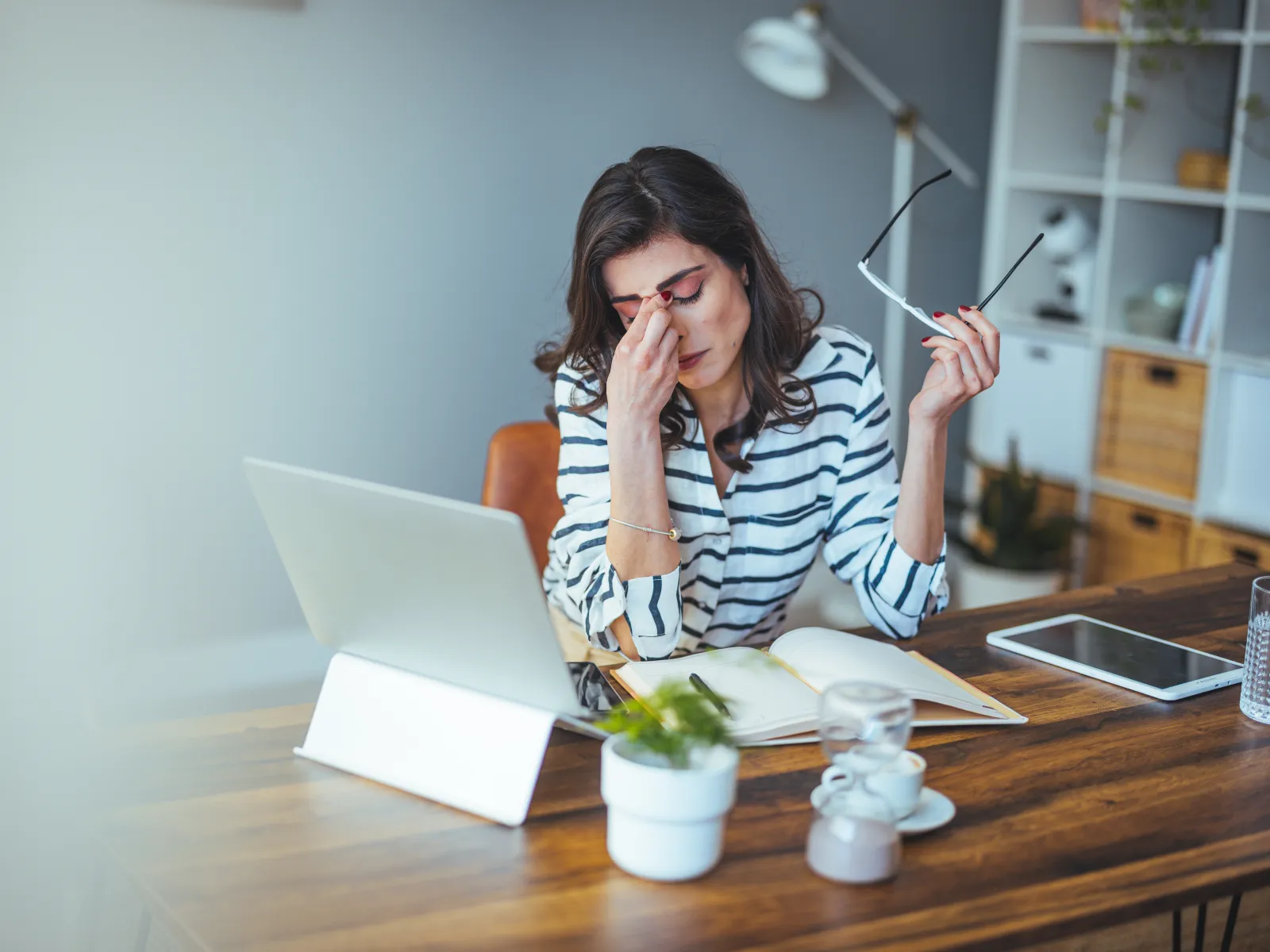 Stressed woman in striped shirt holding glasses and rubbing eyes while working on laptop at wooden desk.