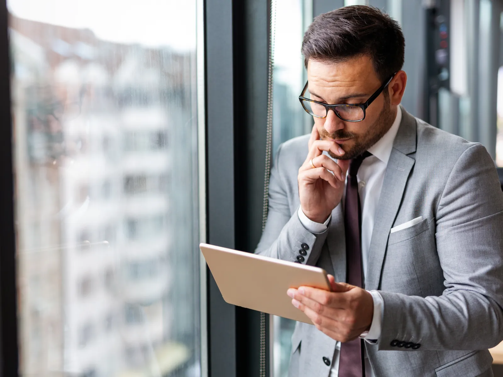Businessman in gray suit and glasses thoughtfully using a tablet by the window in an office.