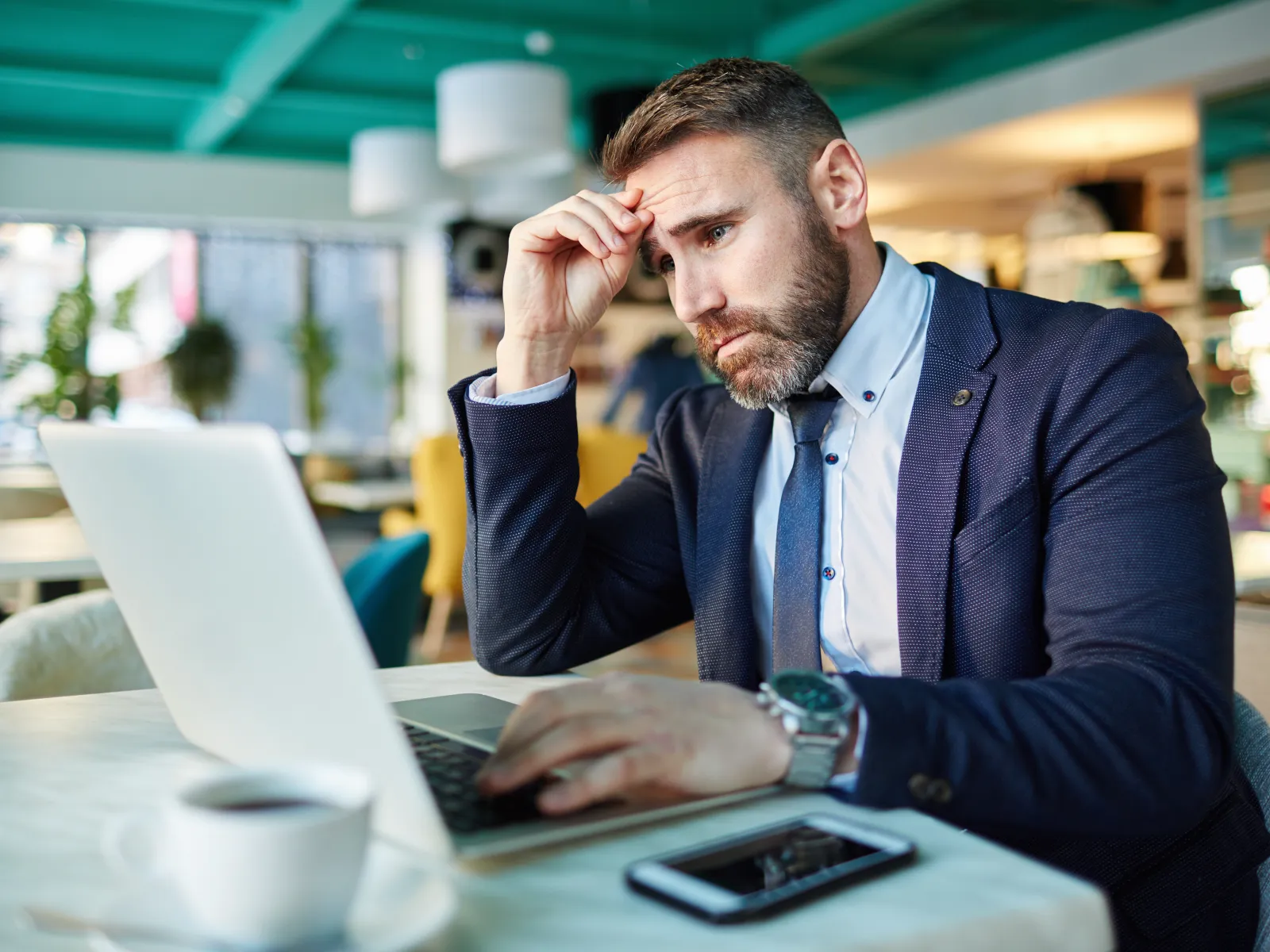 Businessman in suit working on laptop in cafe, looking stressed and focused during work.