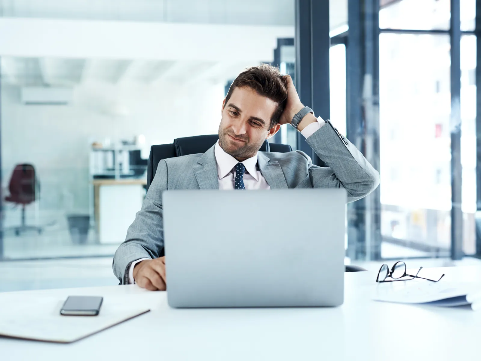 Businessman in gray suit working on laptop in modern office with documents and smartphone on desk