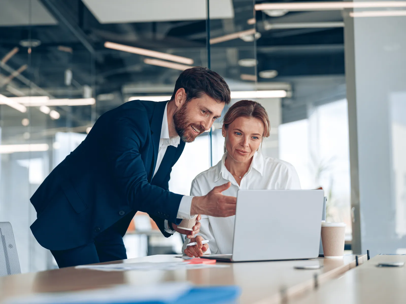 Two colleagues in business attire discussing work while reviewing a laptop at a modern office.