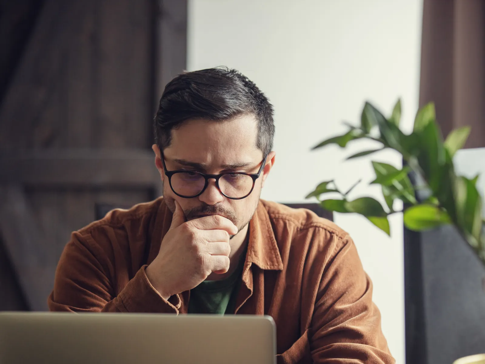 Man with glasses concentrating thoughtfully on laptop screen at home office desk with plant nearby