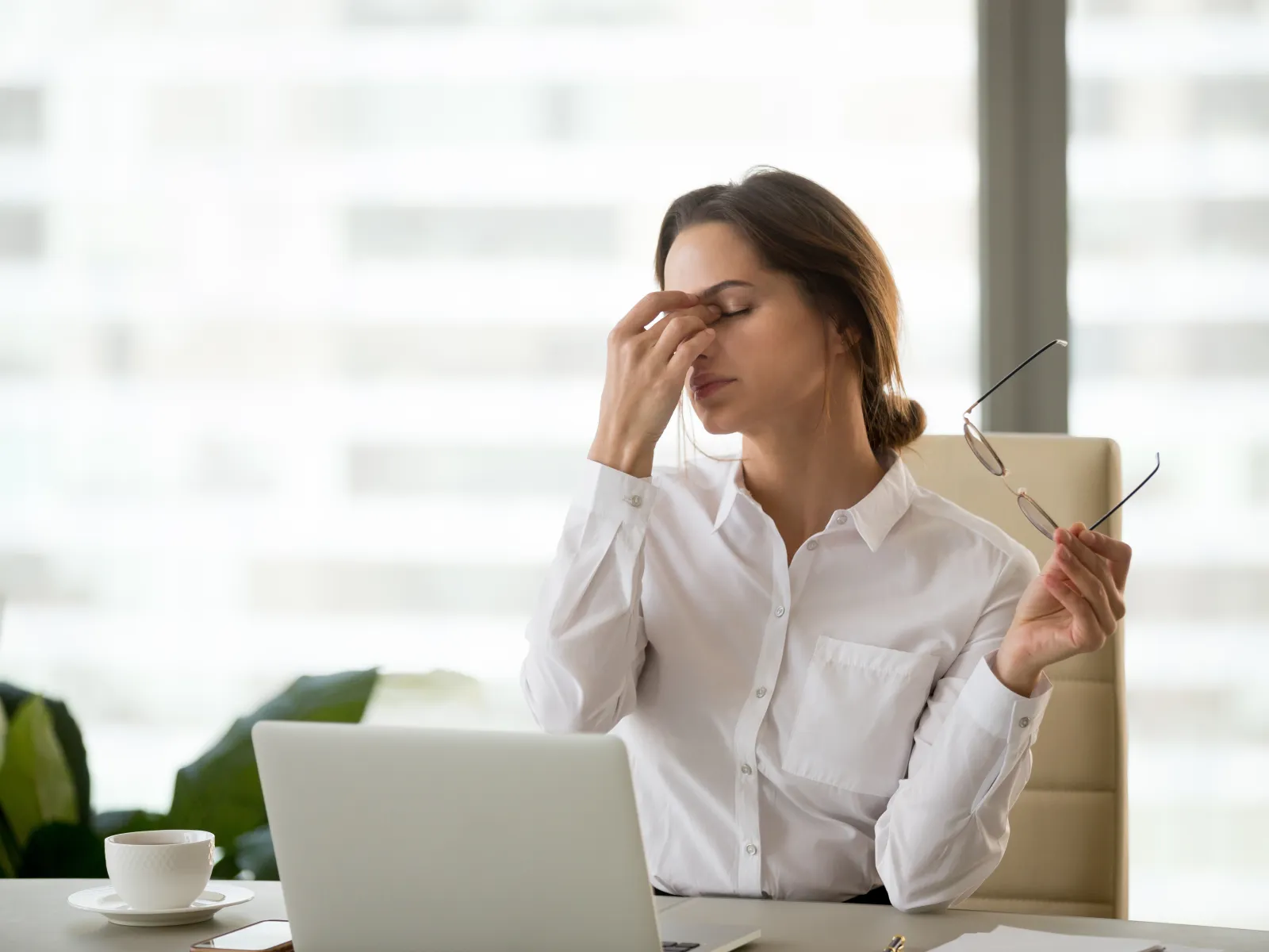 Tired businesswoman in white shirt rubbing eyes while holding glasses at her desk with laptop and coffee cup.