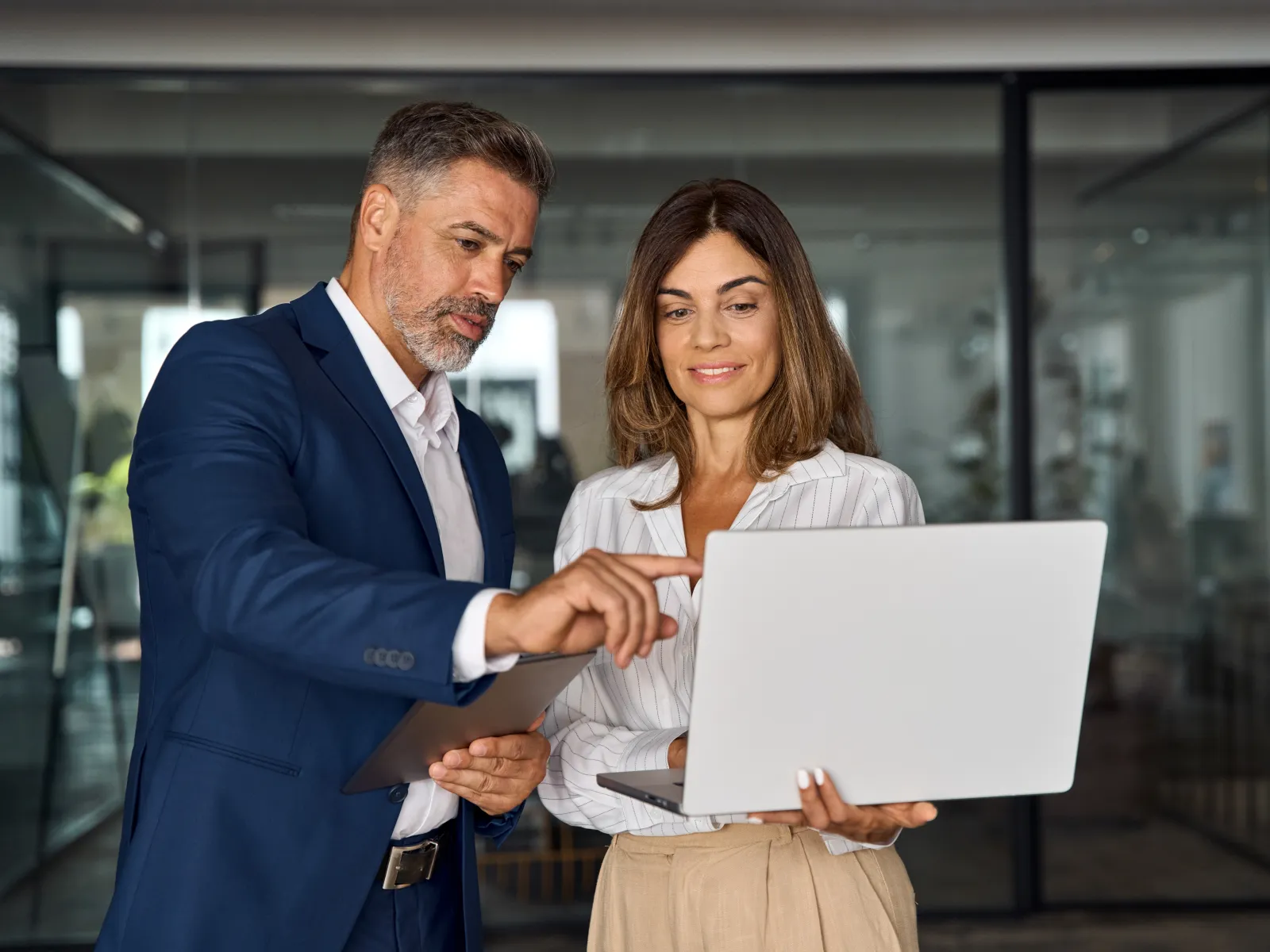 Two business professionals discussing work on a laptop inside a modern office with glass walls.
