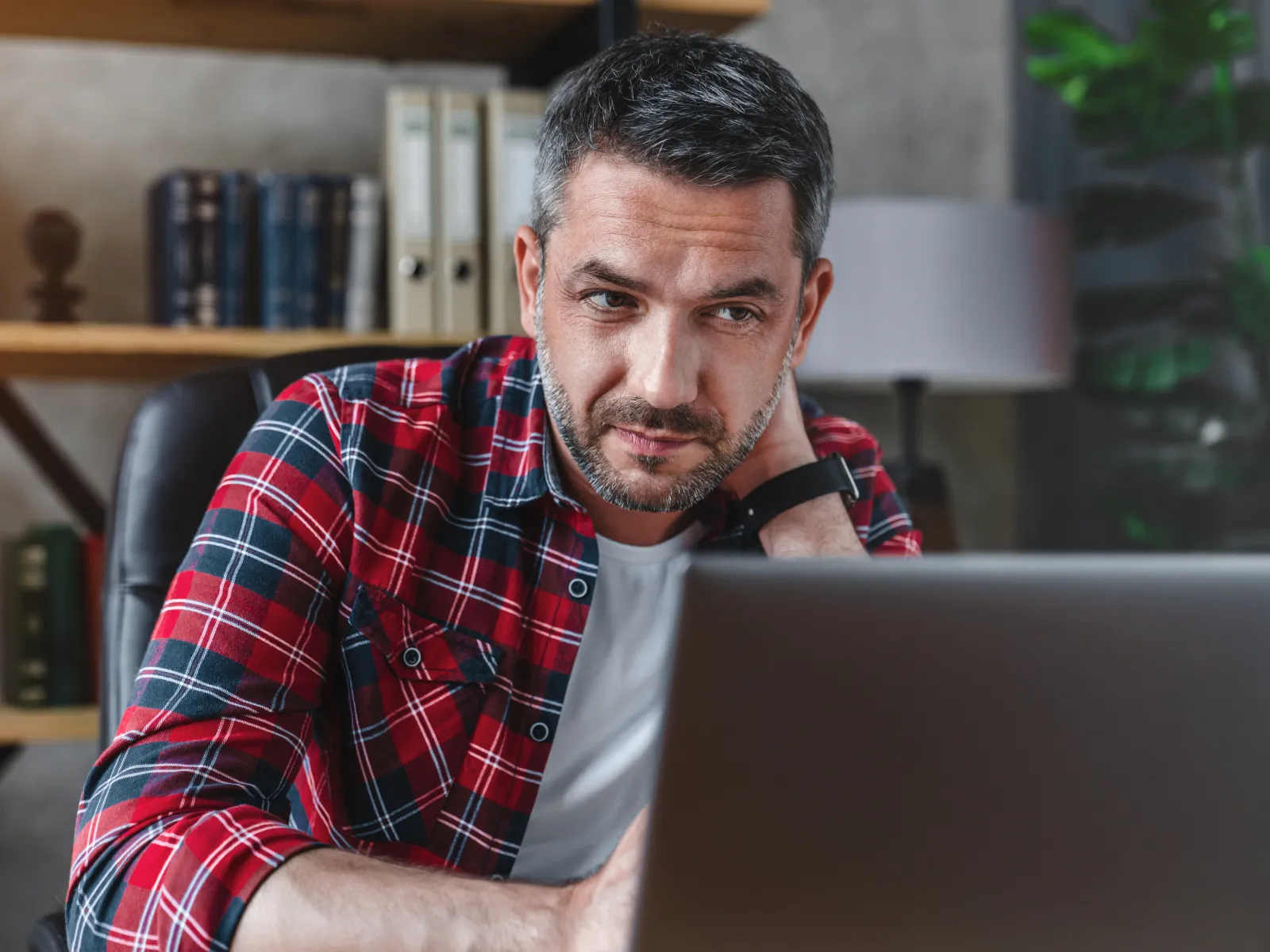 Man in red plaid shirt looking thoughtfully at a laptop screen in a cozy office setting.