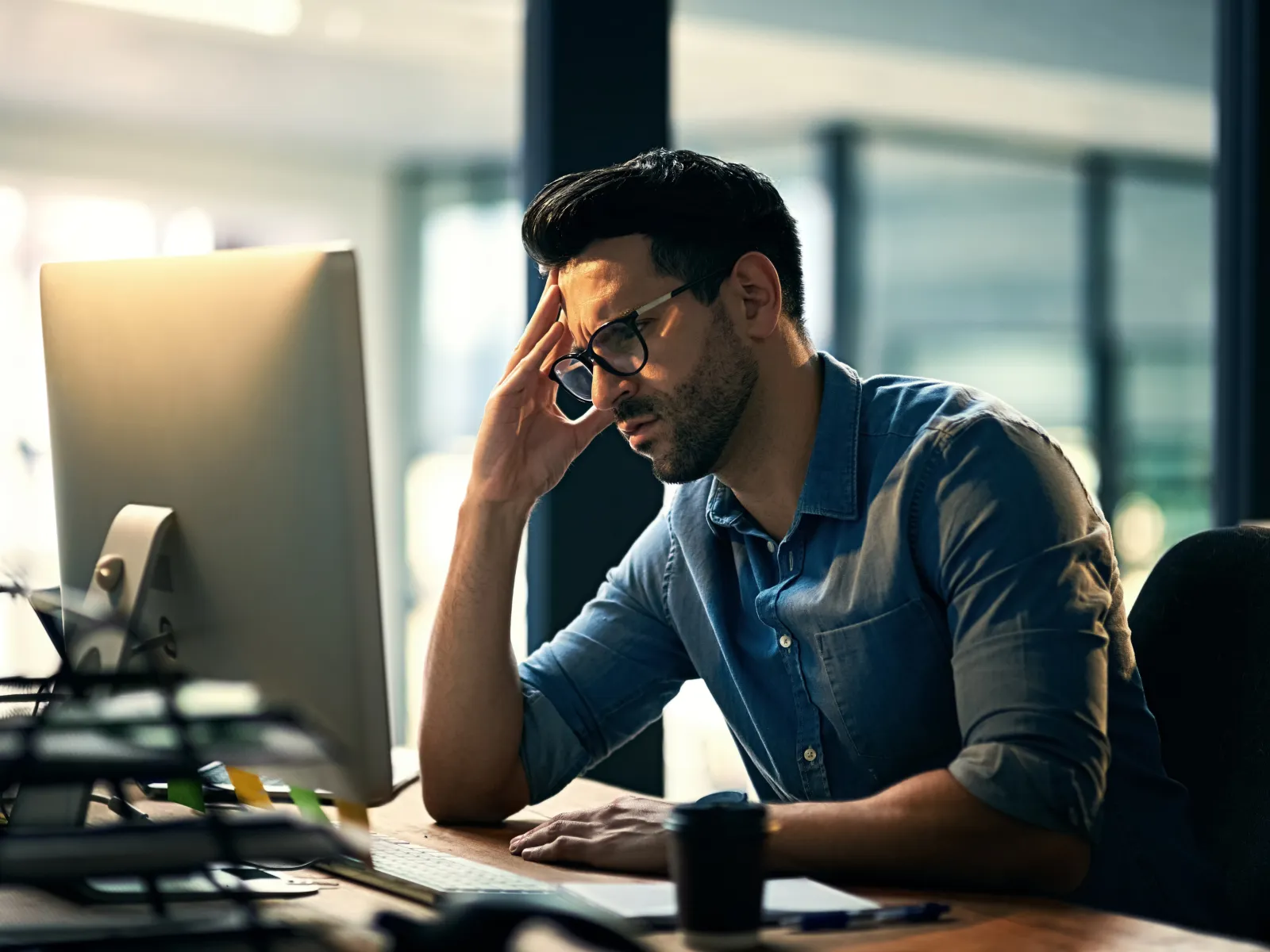 Stressed man in glasses working late on computer in modern office with cup on desk