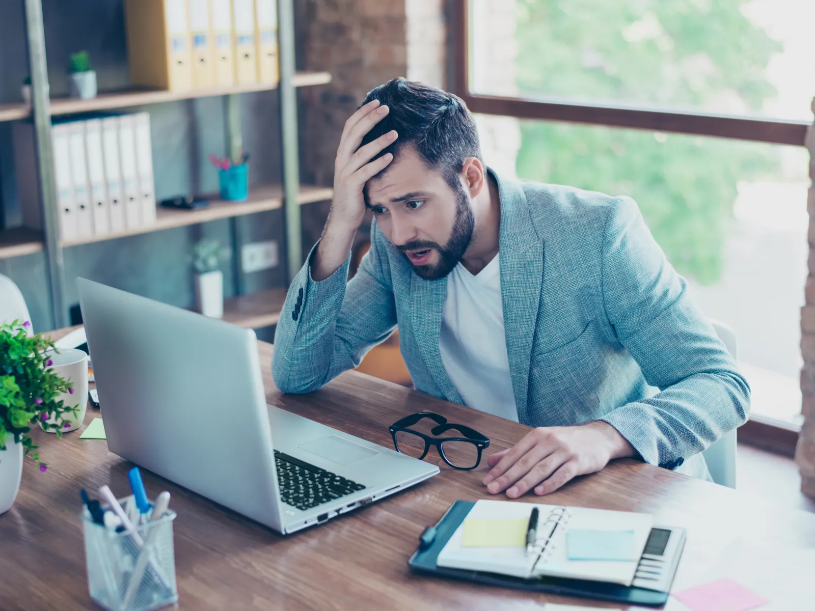 Stressed young man in a gray blazer looking shocked at his laptop screen at a wooden desk in a modern office.