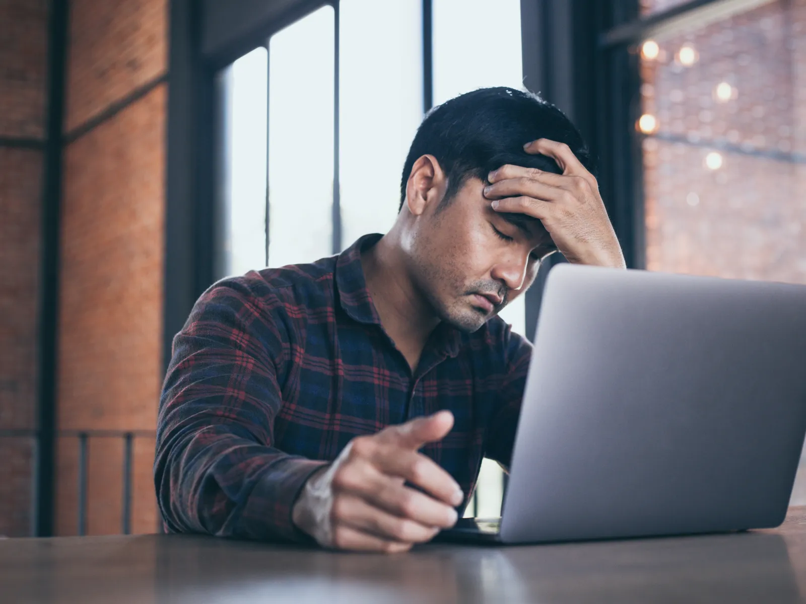 Stressed man sitting at a table with laptop, holding his forehead in frustration in a modern office space.