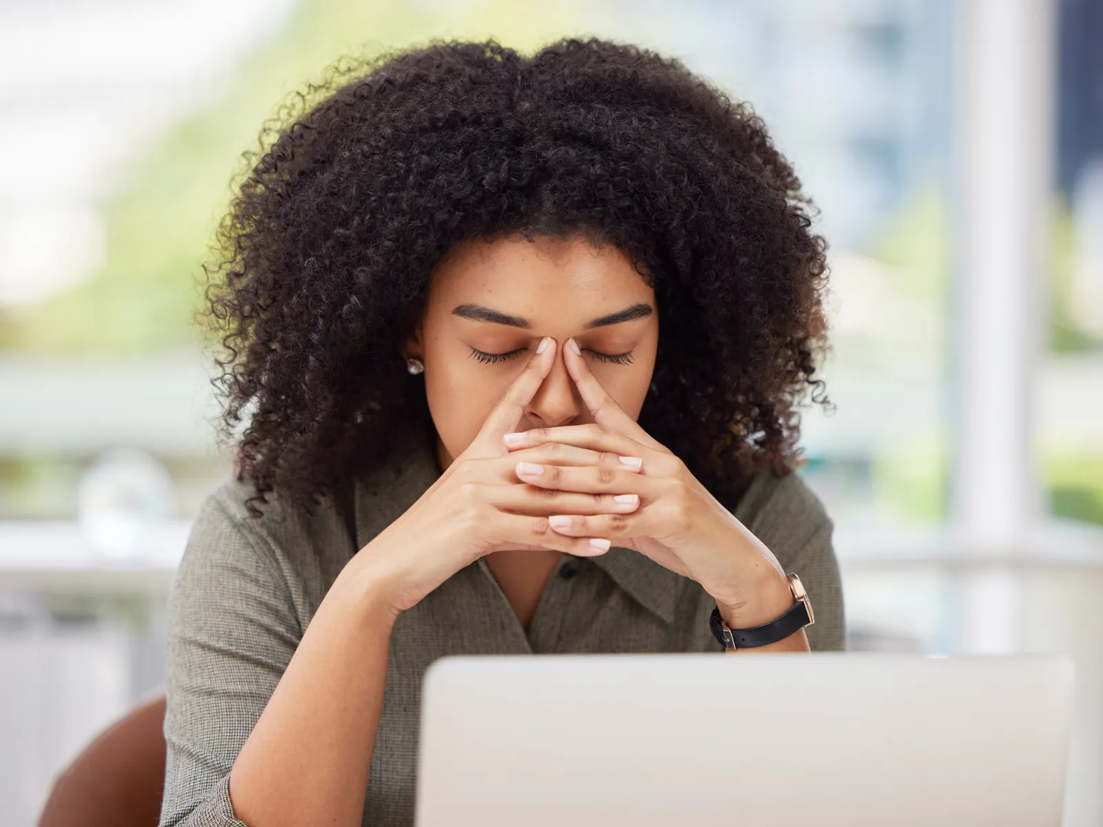 Stressed woman rubbing her eyes while working on a laptop in a bright modern office environment.