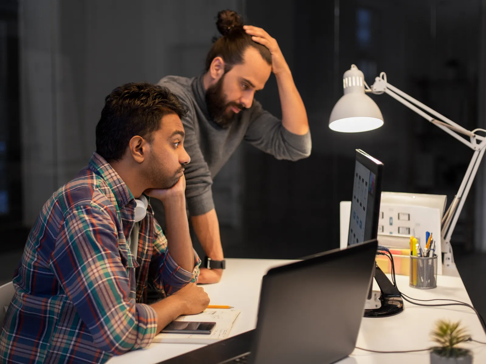 Two men focused and concerned working late at office desk with computers and documents under desk lamp light