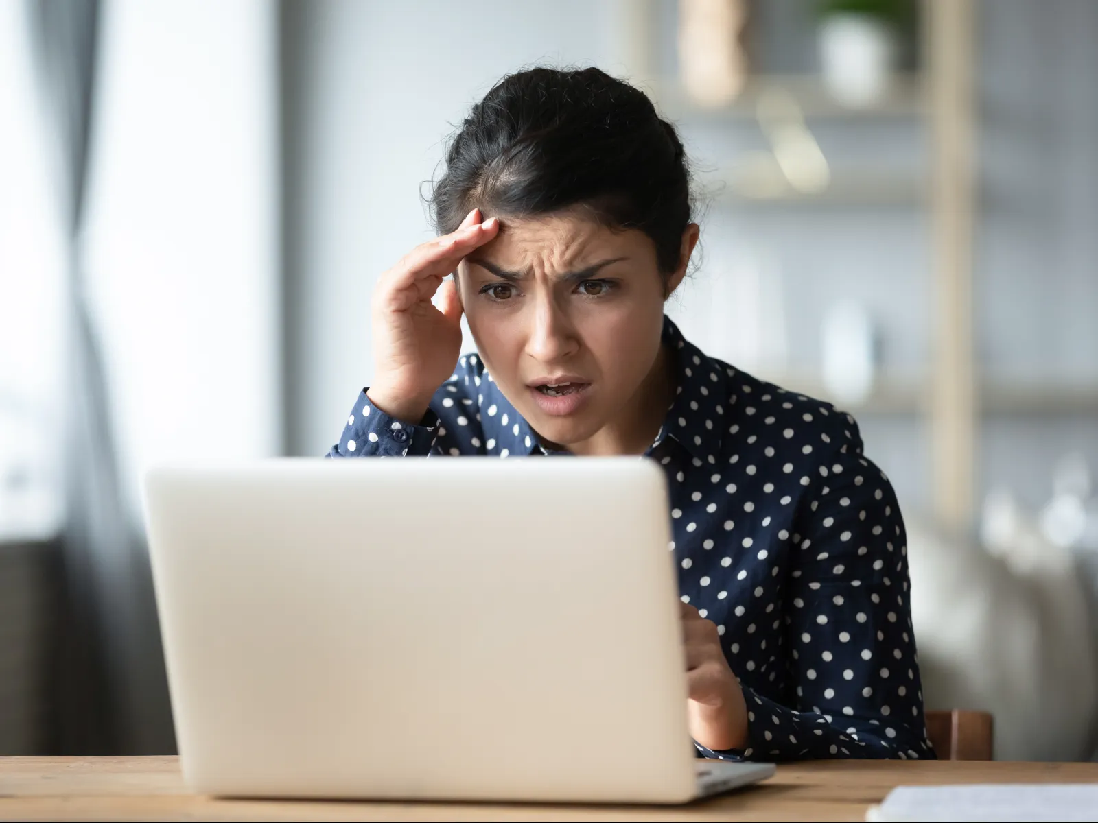 Worried woman in polka dot shirt looking stressed at laptop screen at home desk