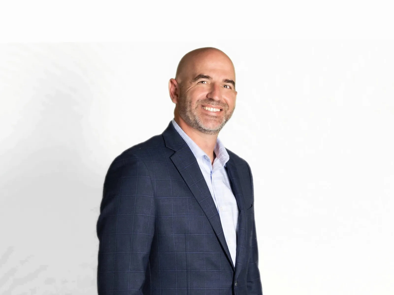 Smiling bald man in a dark blue checked blazer and light blue shirt standing against a white background.
