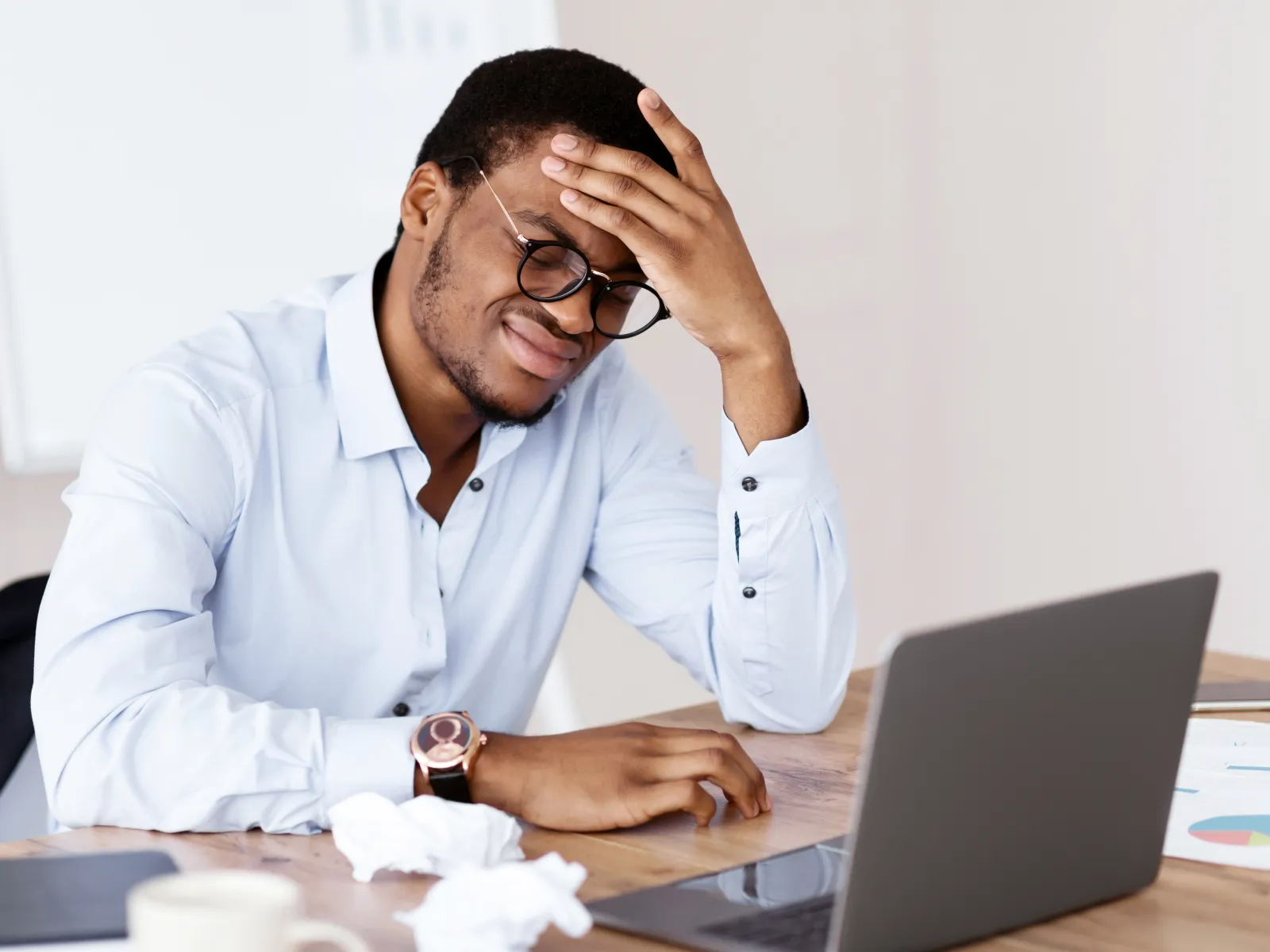 Frustrated man sitting at desk with laptop, holding head in pain, surrounded by crumpled paper and documents.
