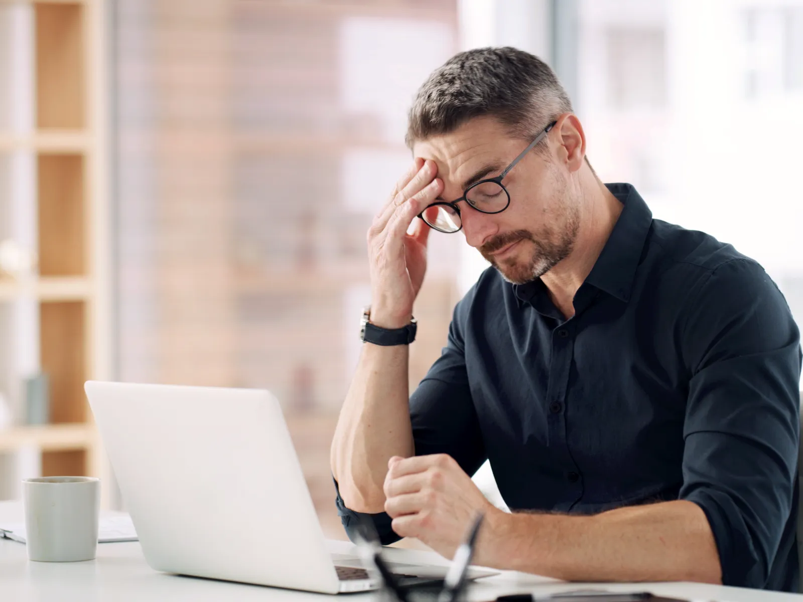 Man in glasses holding head, looking stressed while working on laptop in a bright modern office.