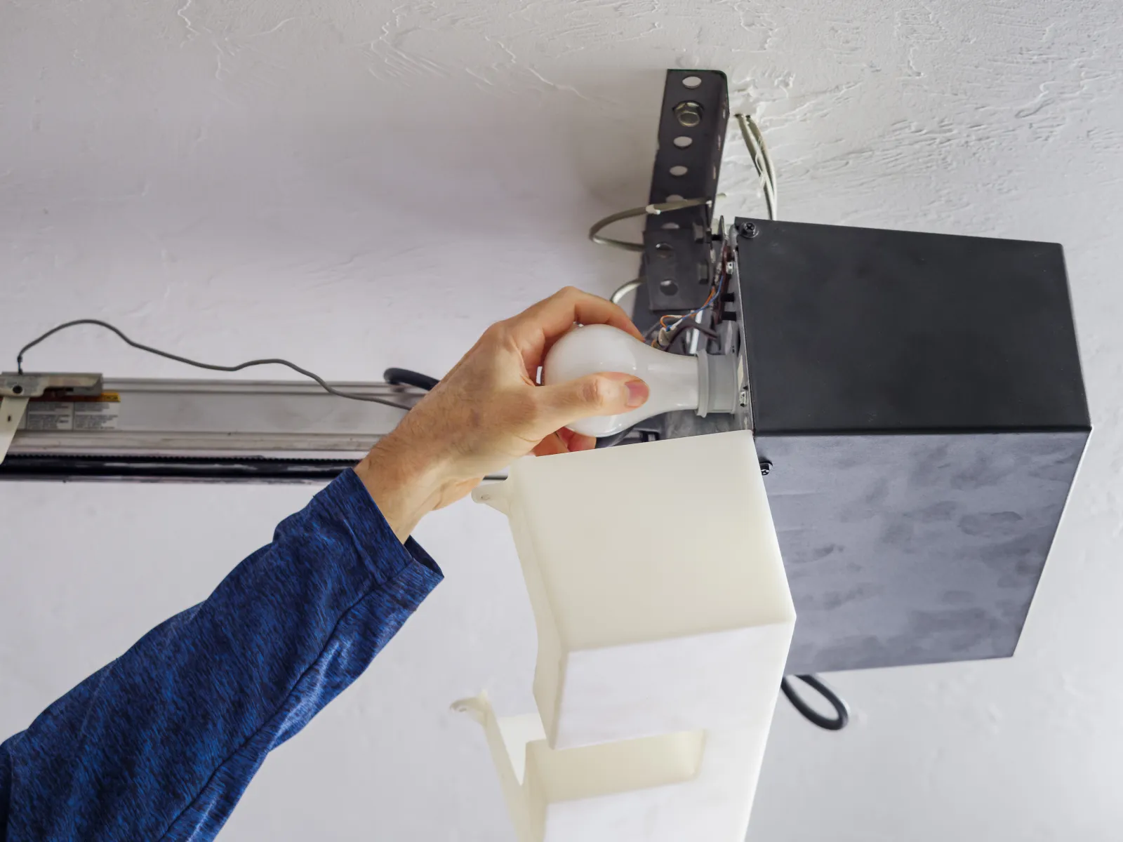 Hand replacing a light bulb in a garage door opener fixture against a white ceiling.