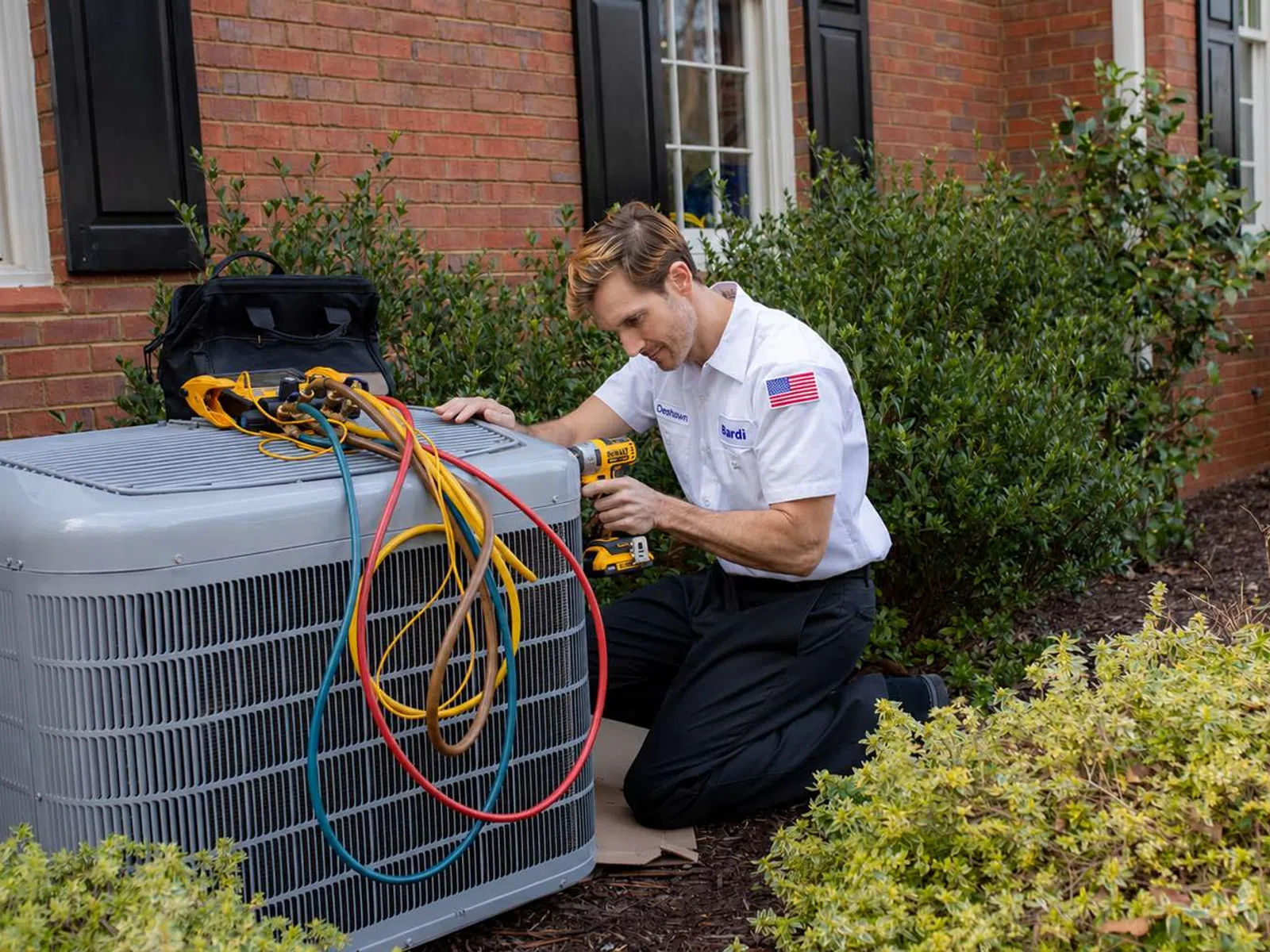 Technician in white uniform repairing outdoor air conditioning unit with colorful cables and power drill beside a brick house.
