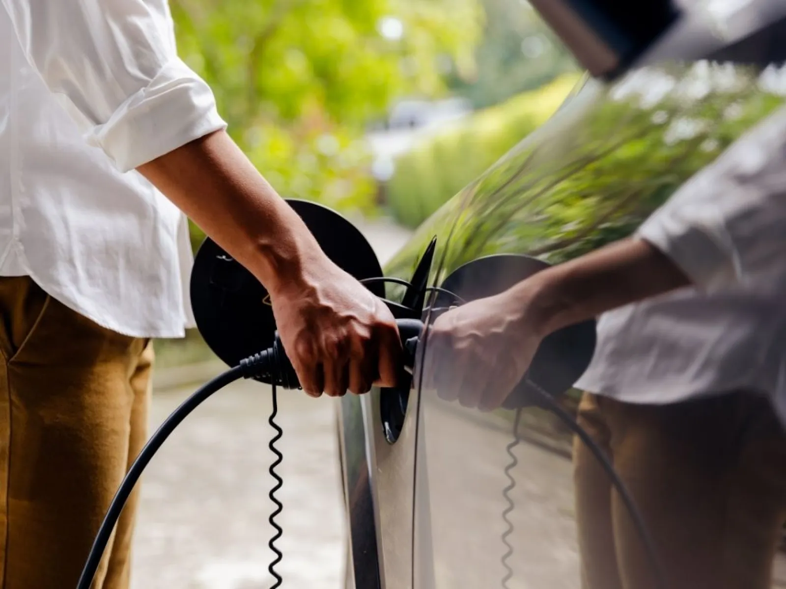 Person plugging a white electric vehicle charger into a white car at a charging station outdoors.