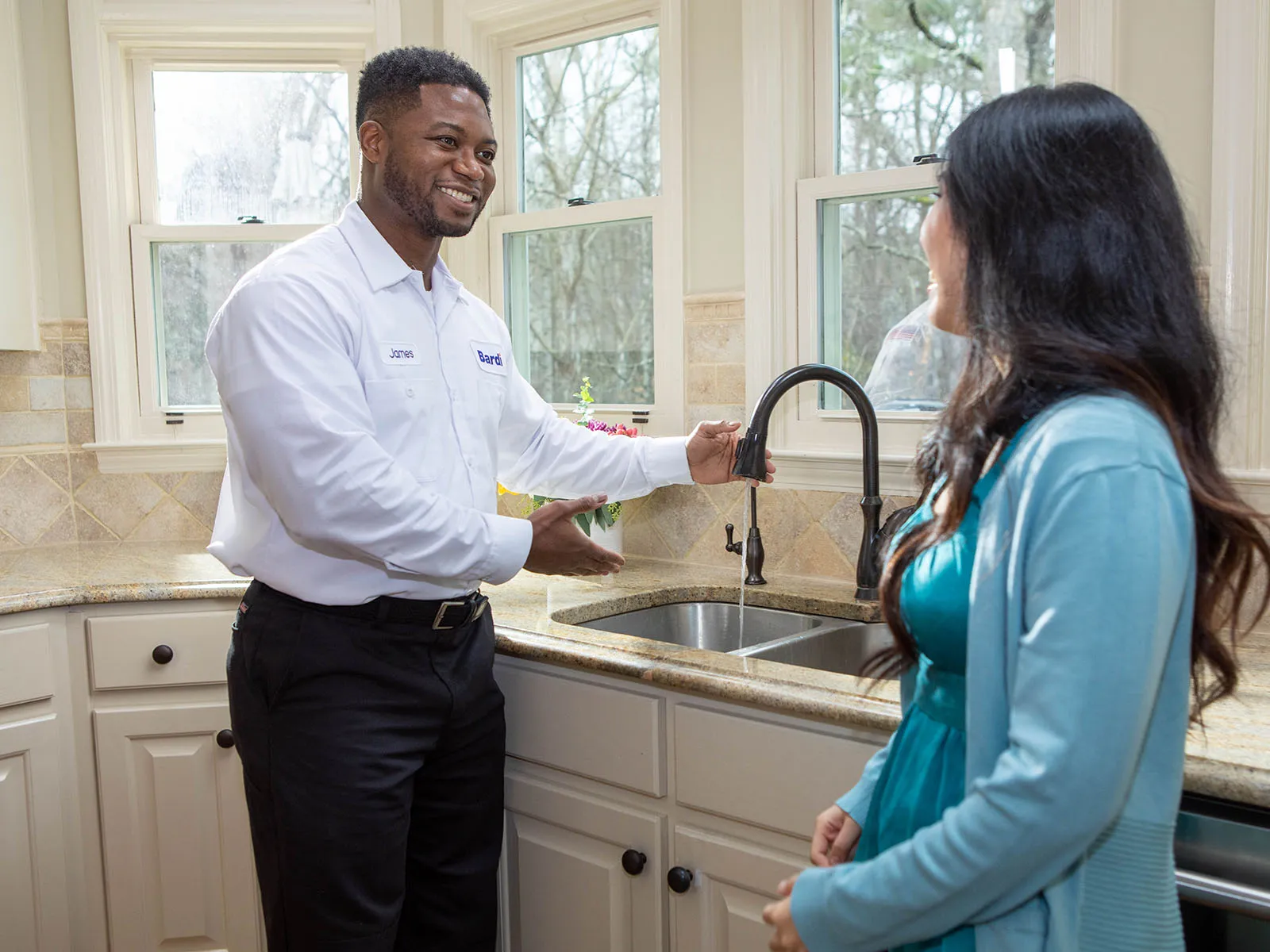 A technician demonstrates a kitchen faucet to a smiling woman in a bright, modern kitchen.