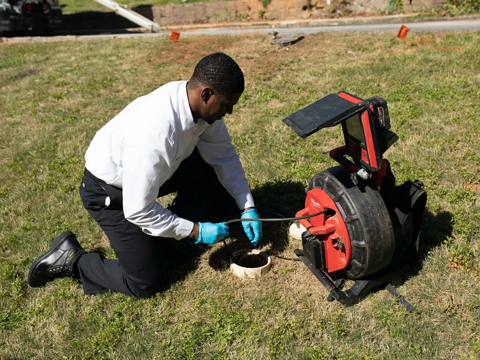 A technician using a plumbing snake at a sewer cleanout on a grassy area.
