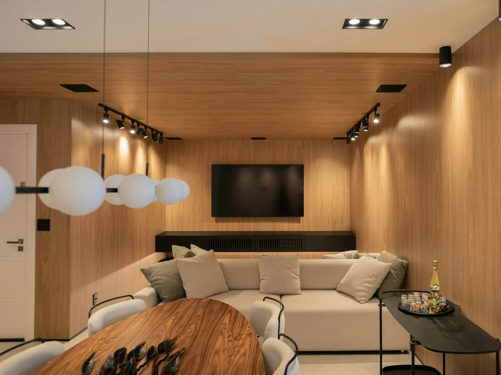 Cozy living room with wood-paneled walls, beige sofa, wooden dining table, black TV, and modern lighting.