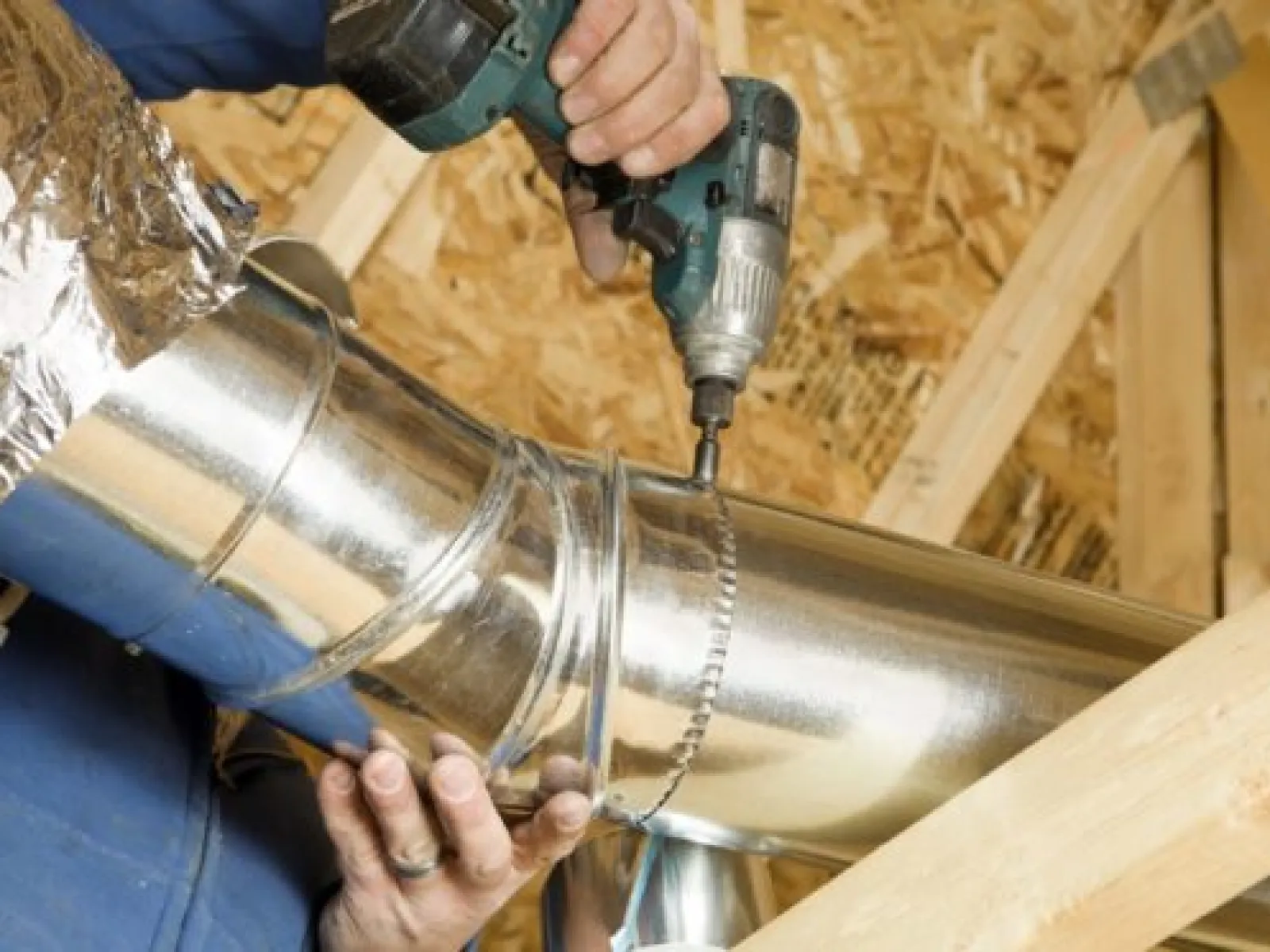 A worker installs a shiny metal duct using a power drill in an attic space.