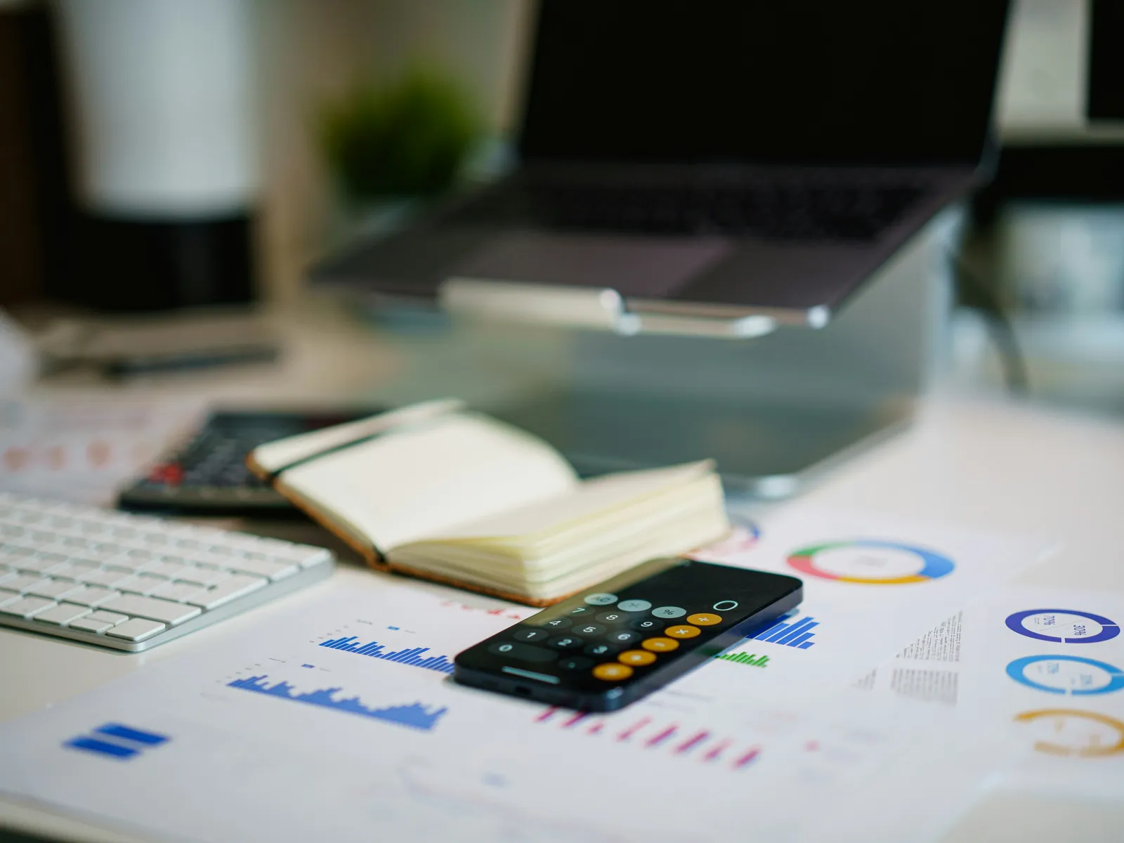 Office desk with laptop, keyboard, calculator, notebook, and financial charts and graphs spread out.
