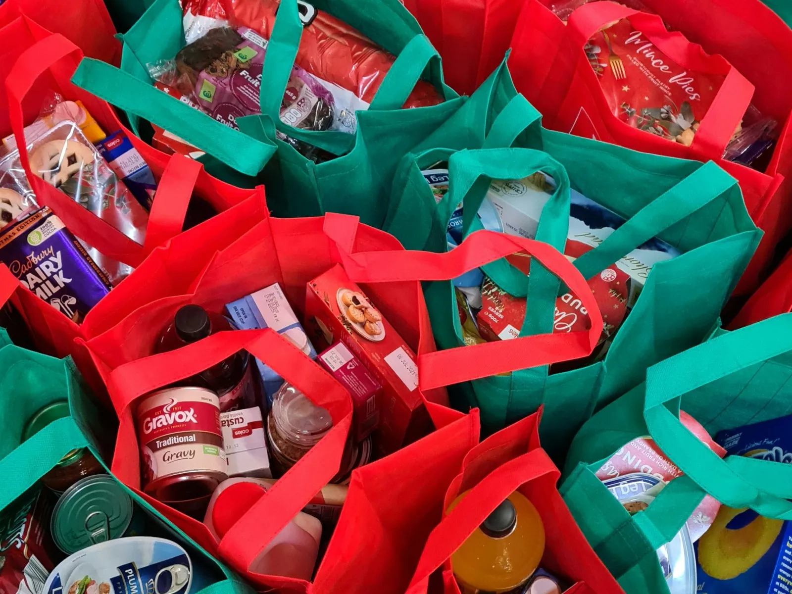 Rows of red and green reusable bags filled with assorted grocery and food items on a dark floor.