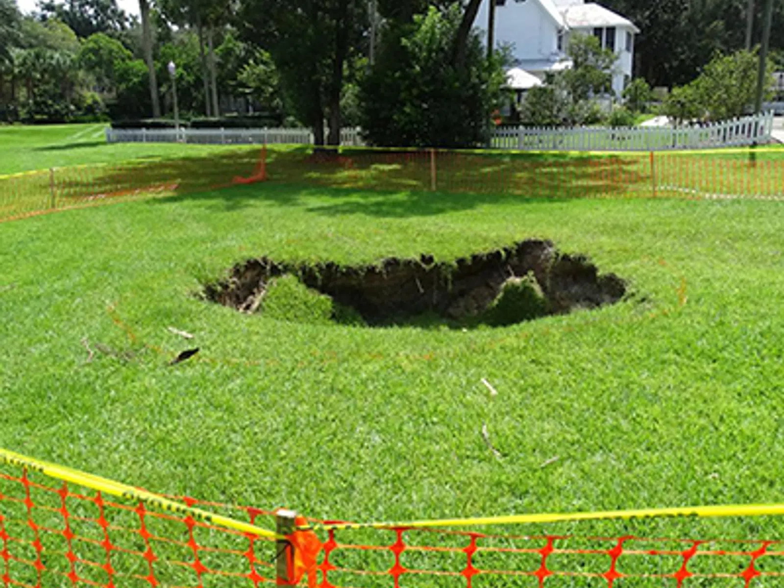 Large sinkhole in a grassy yard surrounded by orange safety fencing near residential houses