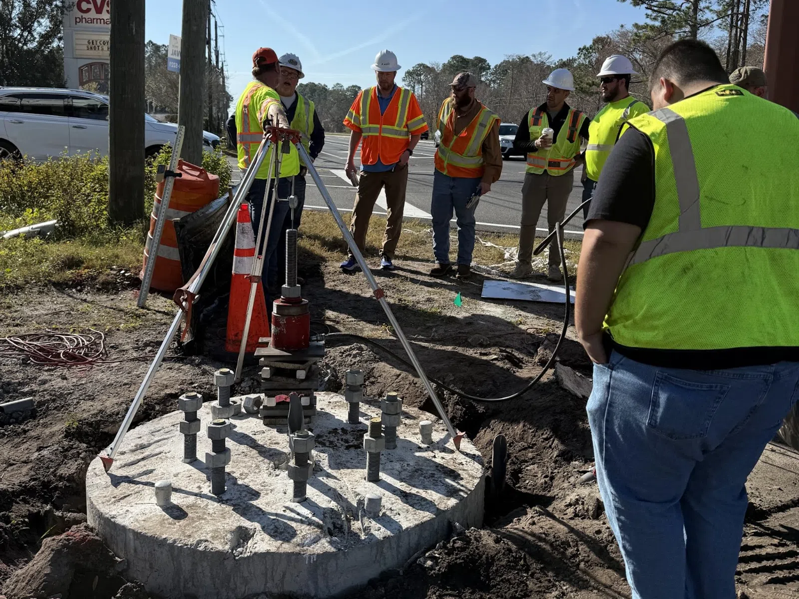 Construction workers in safety vests and helmets inspecting a concrete foundation with bolts by the roadside.