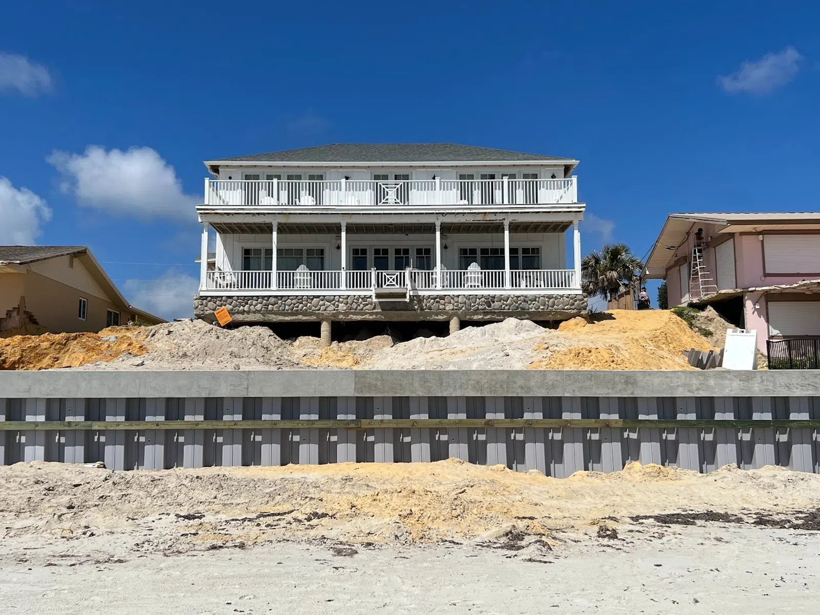 Beachfront house elevated on a sandy dune with a reinforced seawall under a clear blue sky.