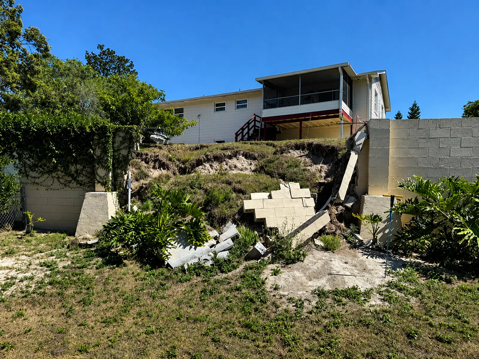 Collapsed retaining wall near a backyard with visible erosion under a residential house on a sunny day
