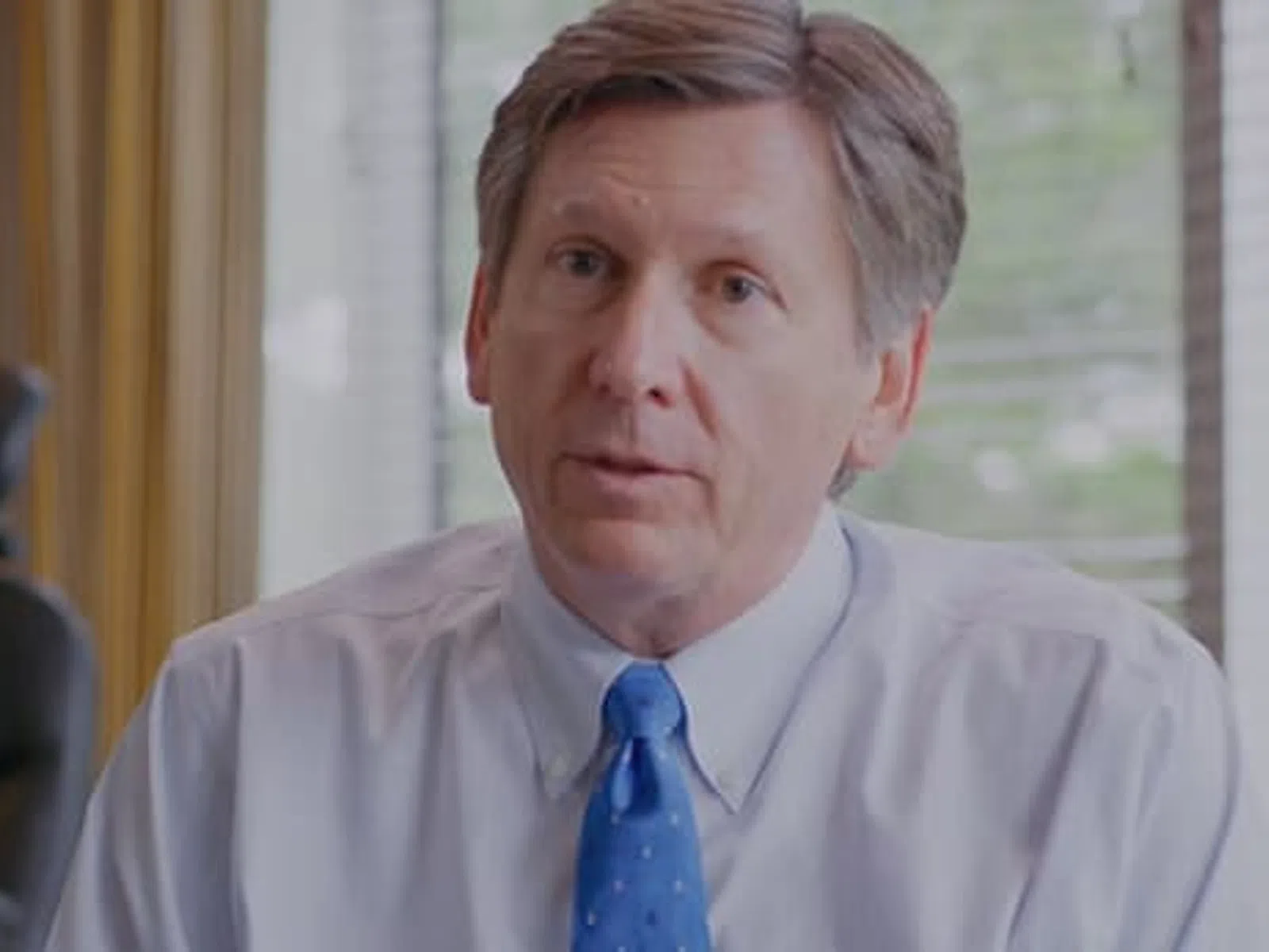 Middle-aged man in office wearing white shirt and blue tie, speaking with serious expression near window blinds.