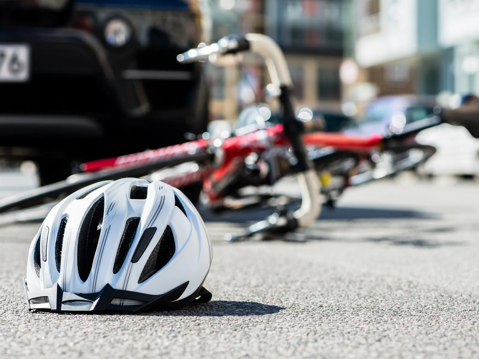 White bicycle helmet on asphalt with a fallen red bicycle and blurred car in the background in daylight.