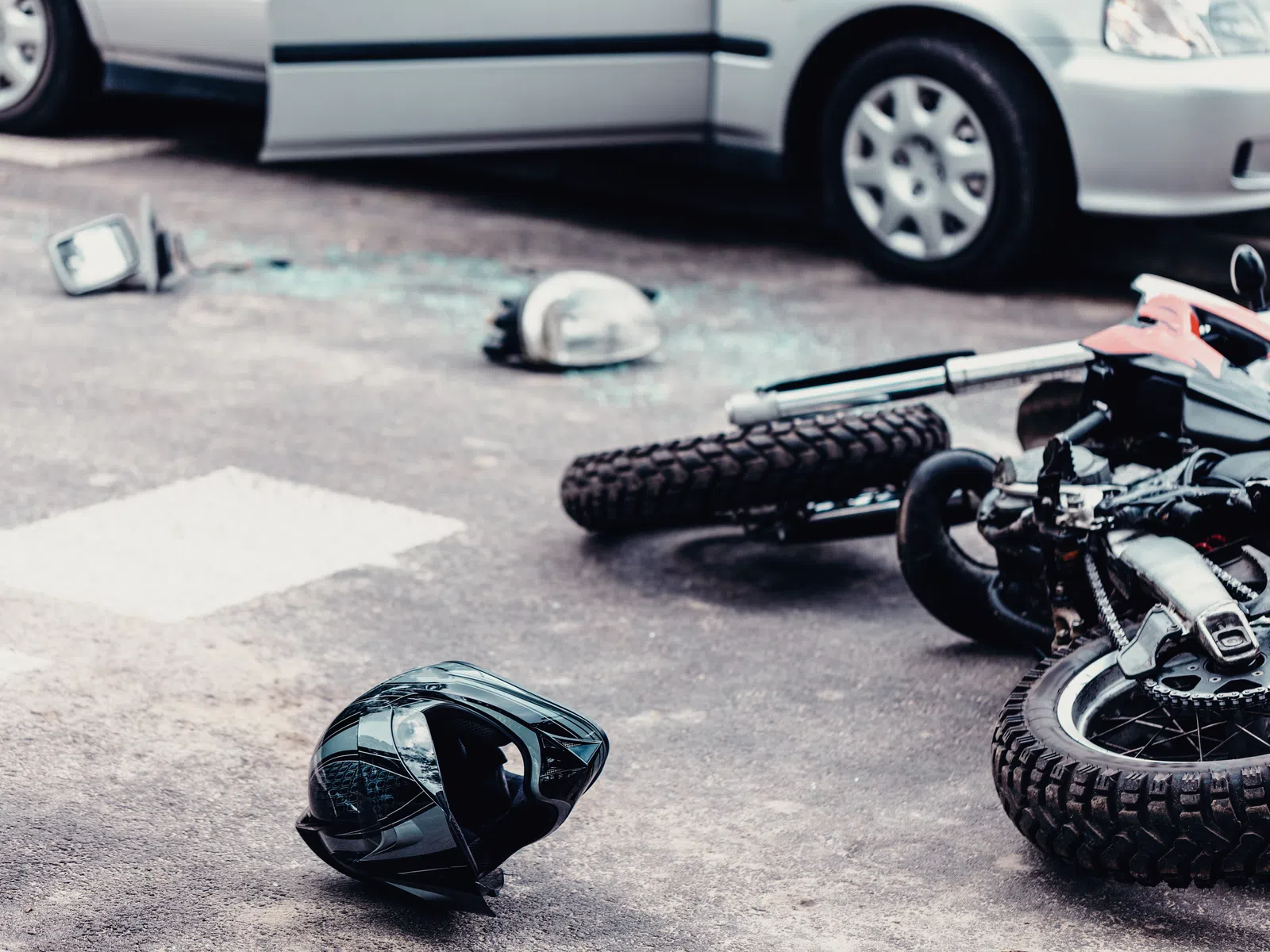 Fallen motorcycle and helmet on the road after a collision with a car in the background and shattered glass nearby.