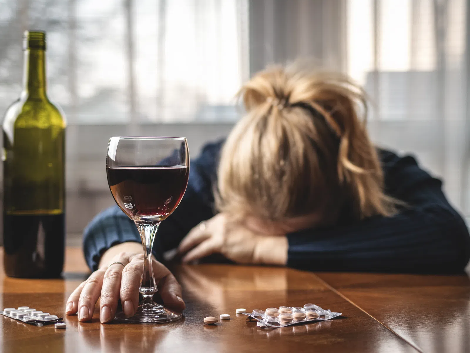 Person with head down at table with wine, pills, and a bottle suggesting distress or substance use.
