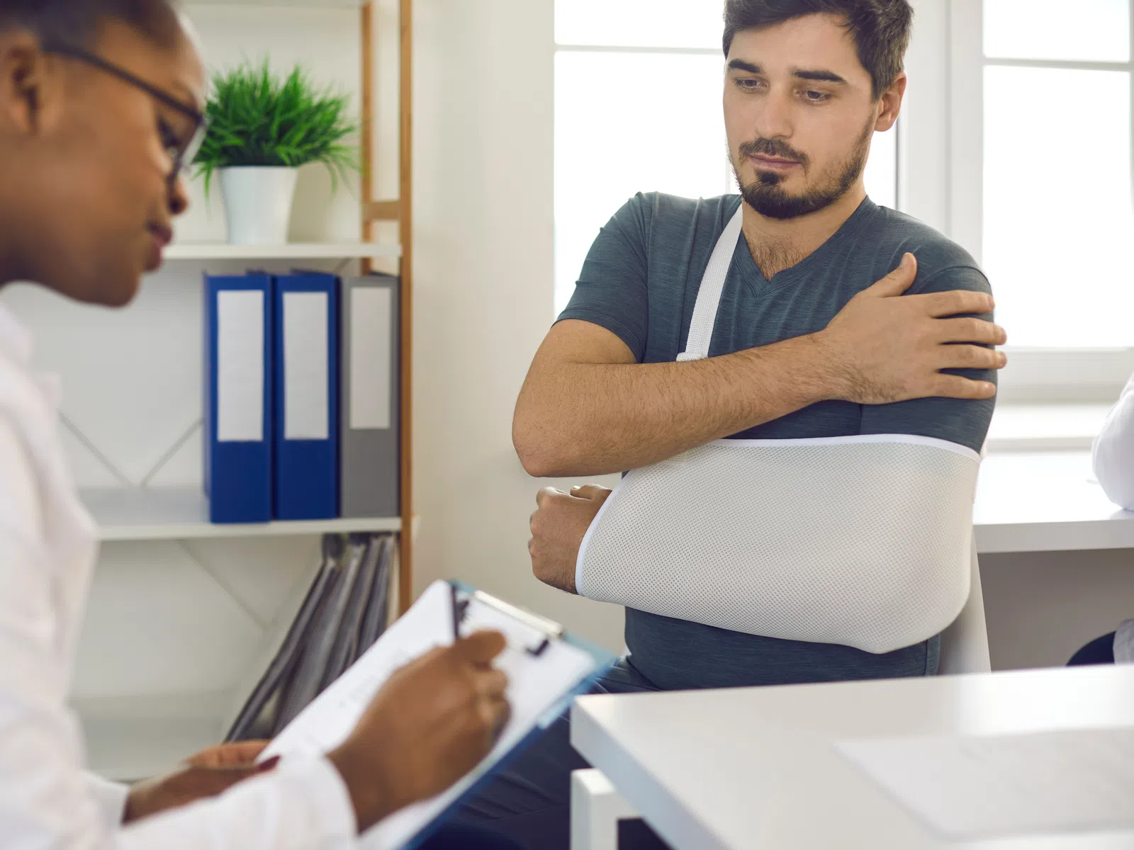 Man with arm in sling consulting a female doctor taking notes in a medical office setting.