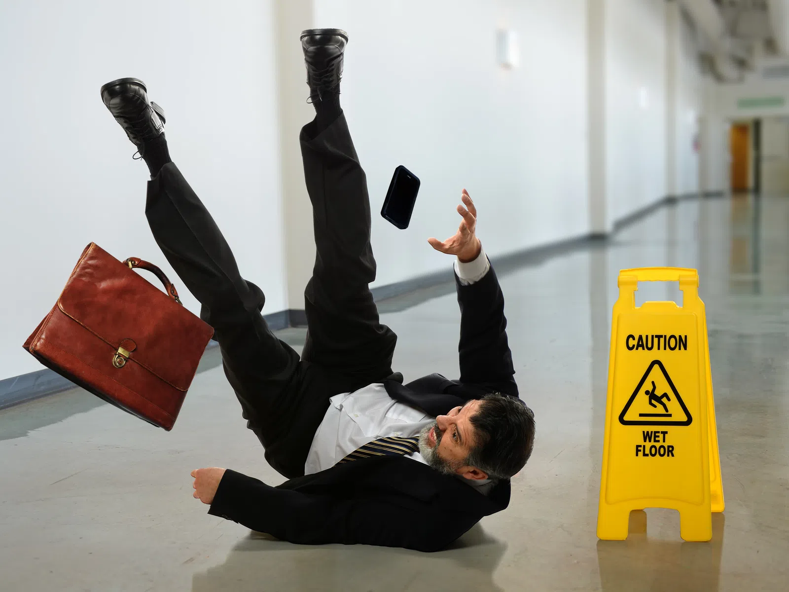 Businessman in suit slipping on a wet floor with briefcase and phone falling beside caution sign in hallway