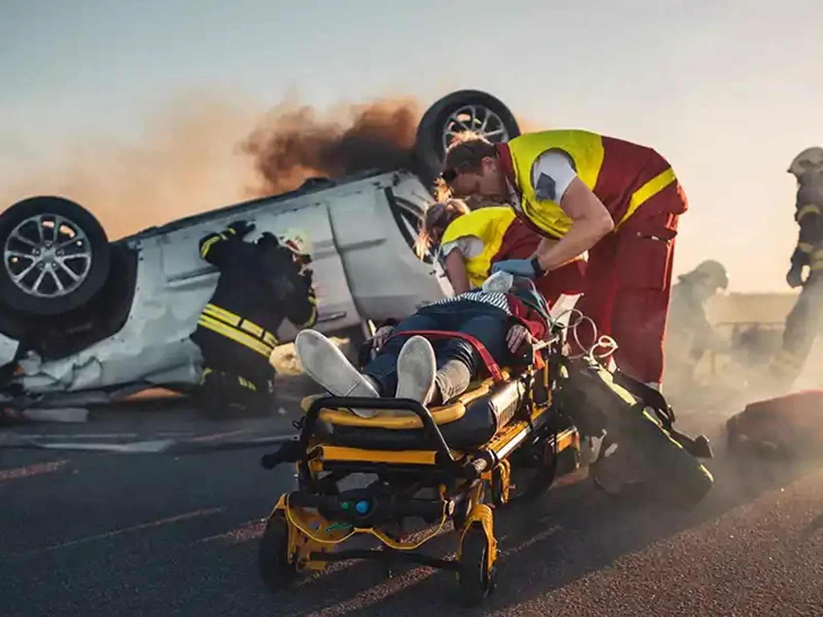 Emergency responders assist injured person beside overturned car emitting smoke during sunset on highway.