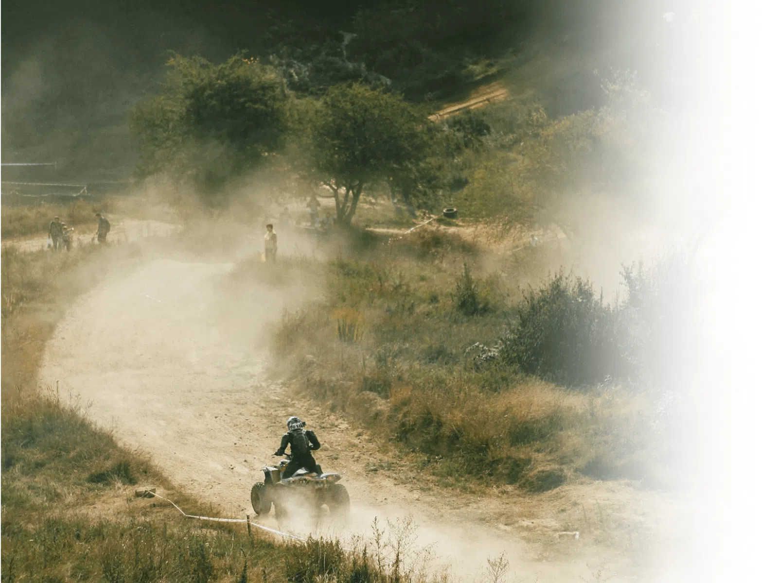Person riding an ATV on a dusty dirt trail surrounded by trees and dry grass in a natural landscape.