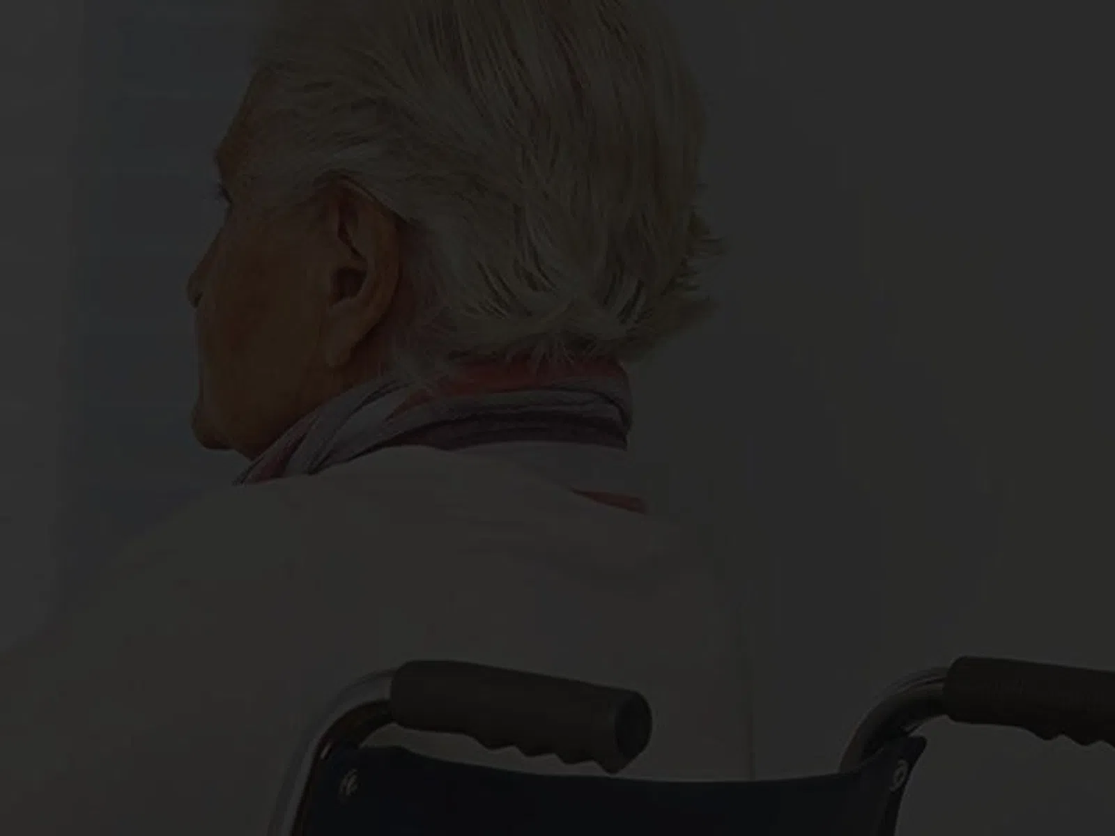 Elderly person sitting in a wheelchair with a sad posture in a dimly lit room near a window.