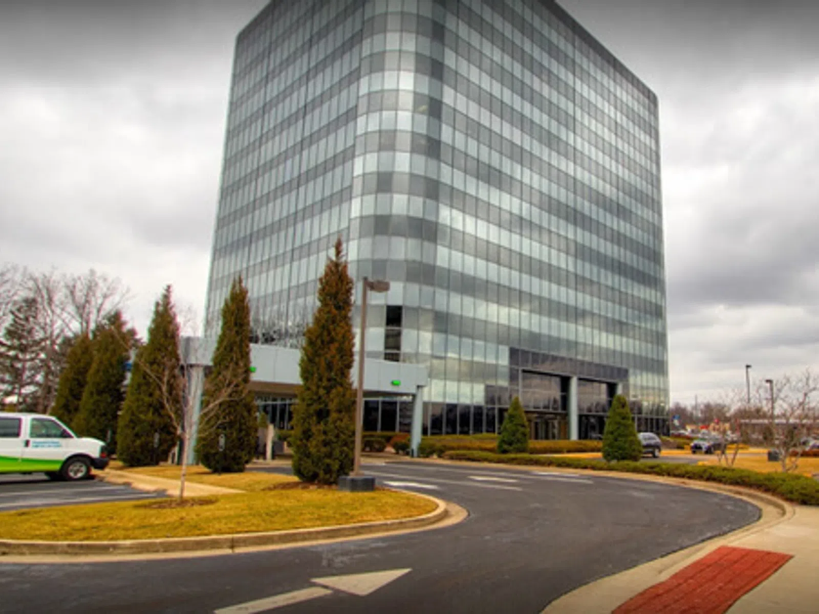 Glass office building with curved driveway, landscaped greenery, and overcast sky in an urban area.