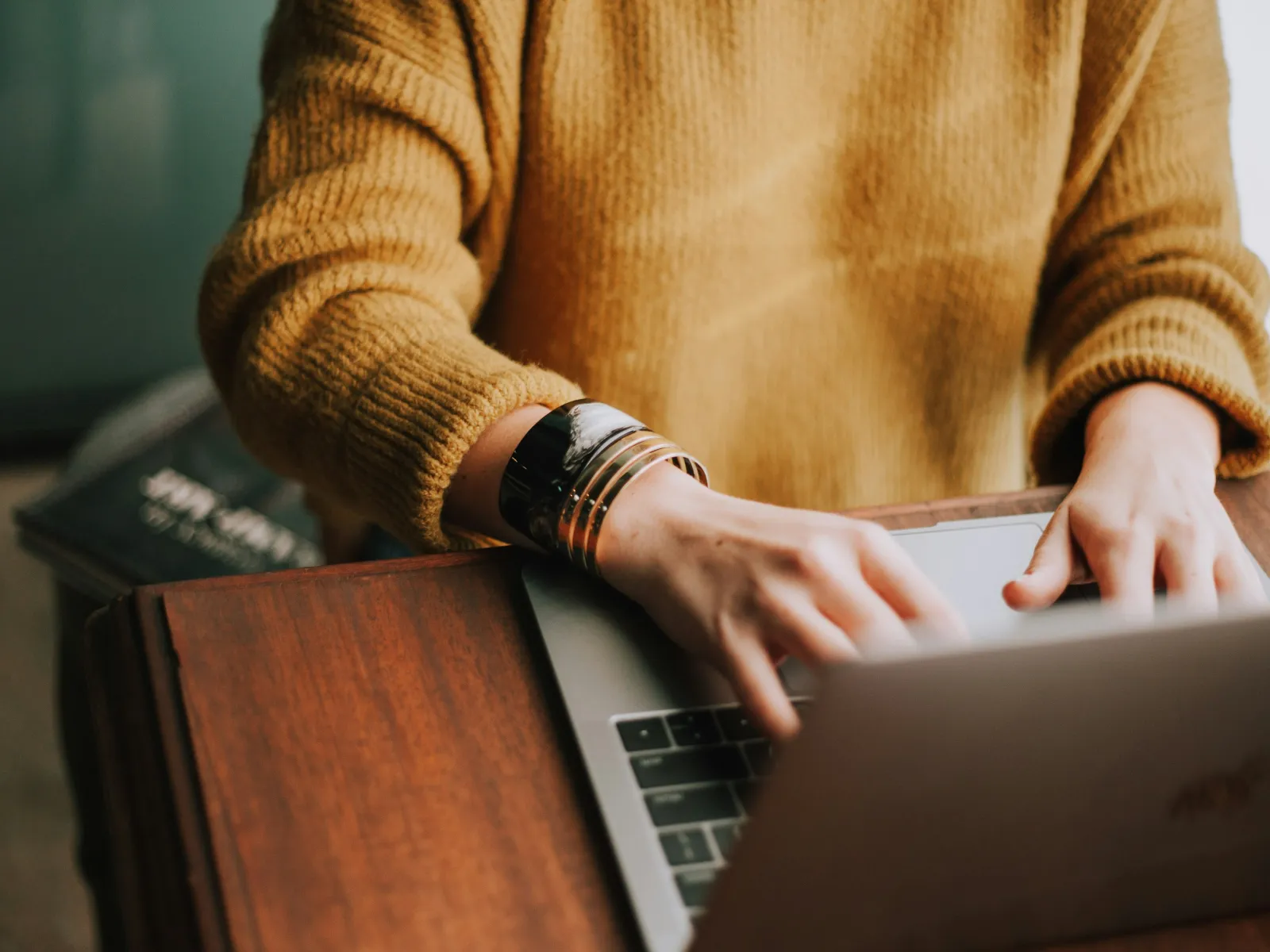 Person wearing a mustard sweater typing on a laptop keyboard at a wooden desk.