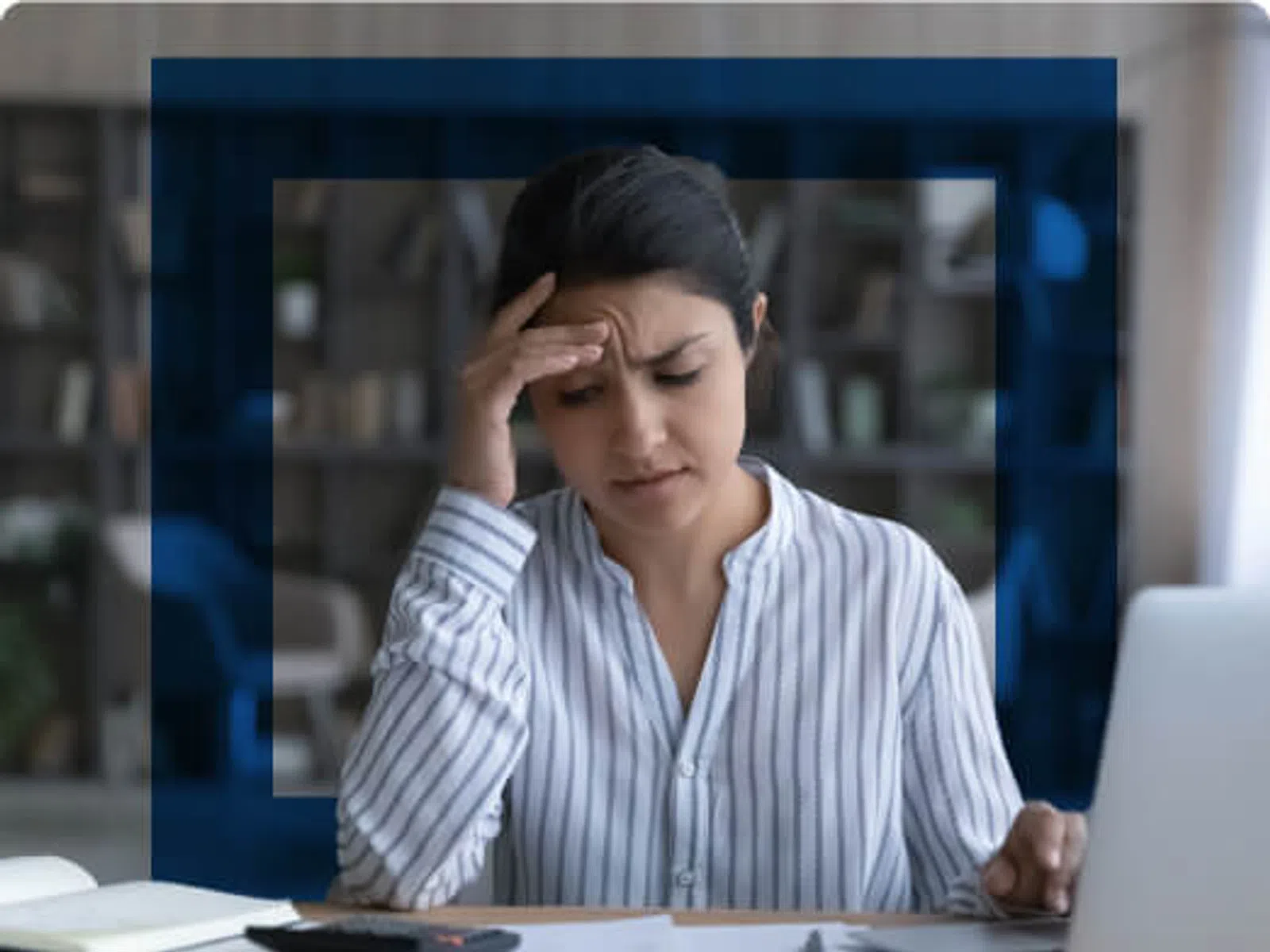 Woman with striped shirt looking stressed while working at a desk with laptop, documents, and calculator.