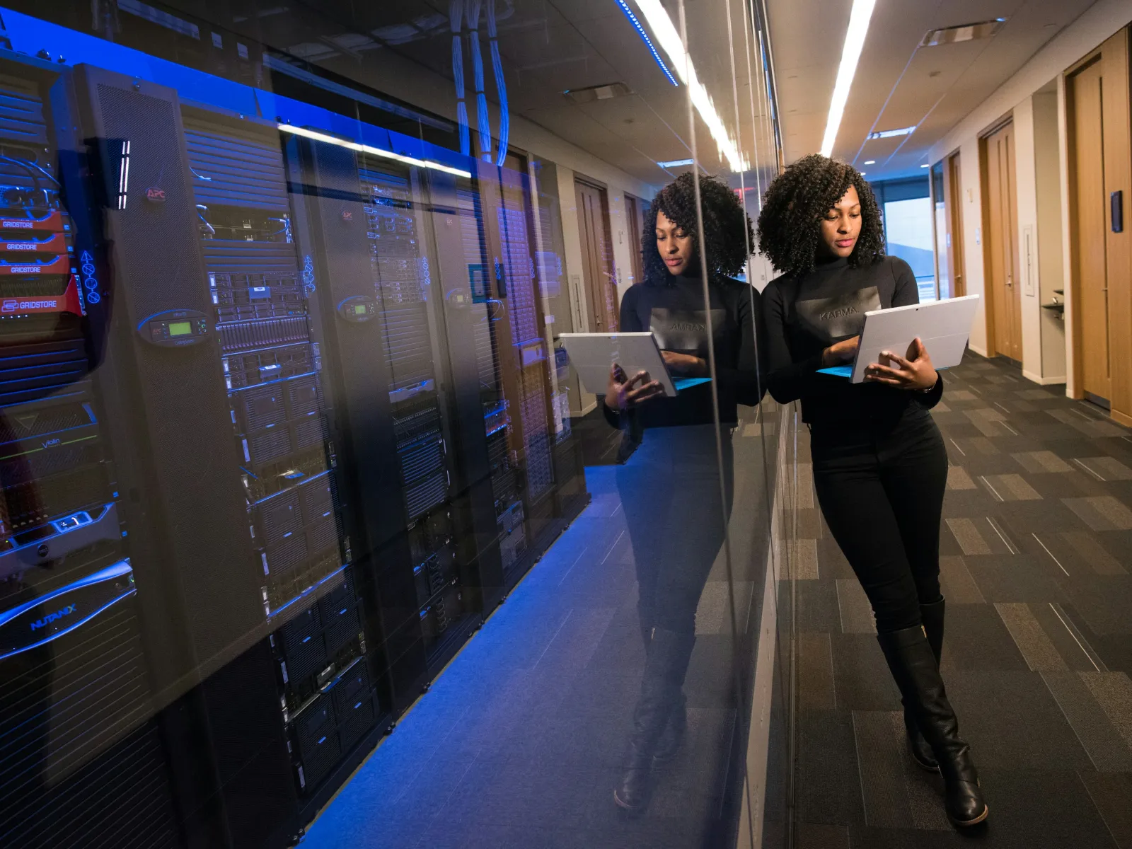 Woman using laptop near server racks in a data center hallway with reflective glass wall.