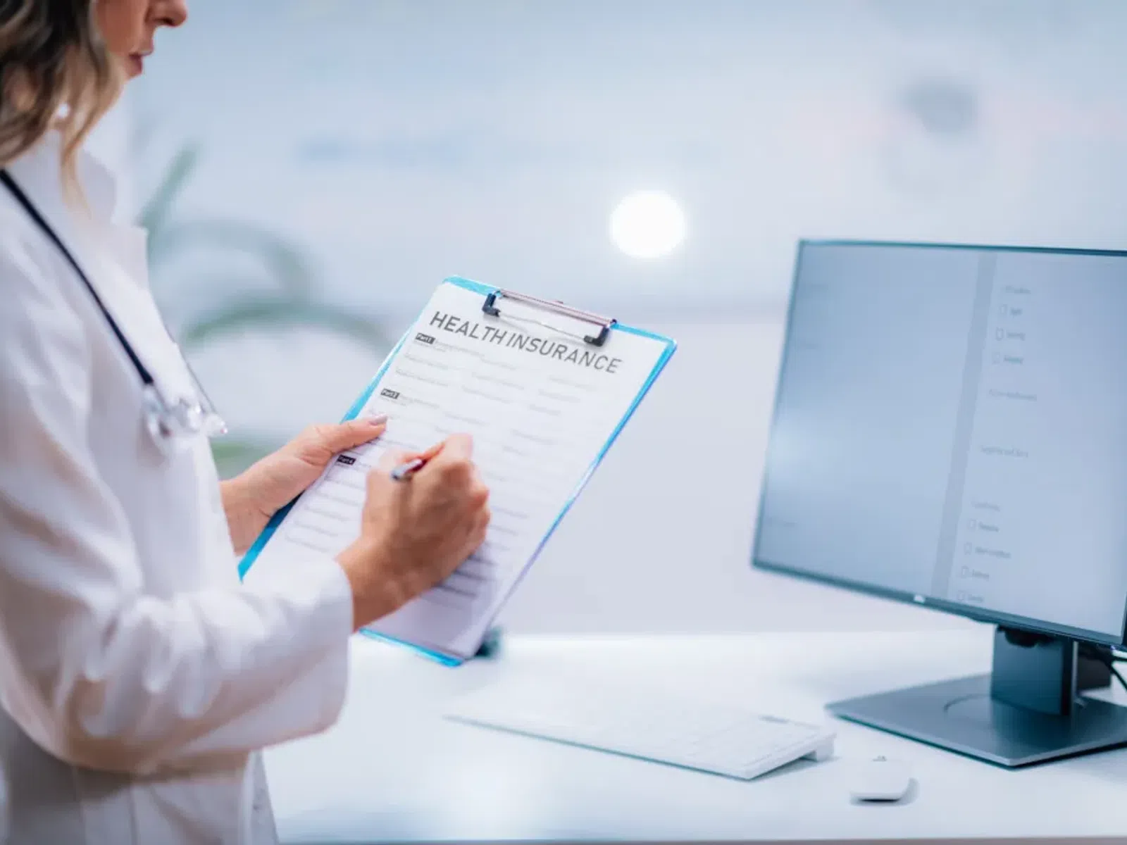 Doctor filling out a health insurance form on a clipboard next to a computer in a medical office.