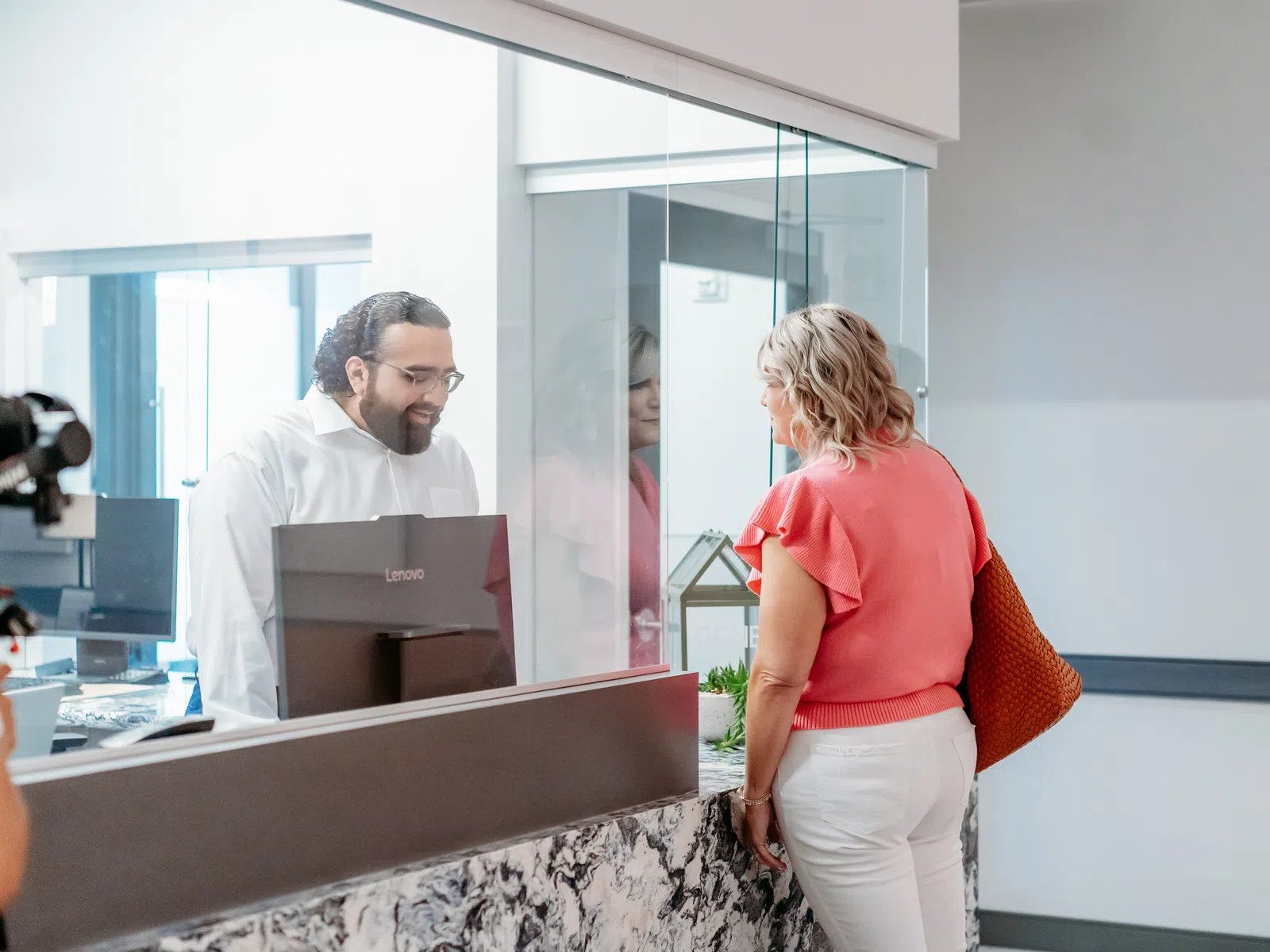 Woman in coral blouse talks to man behind glass desk in modern office with laptop and bright window.