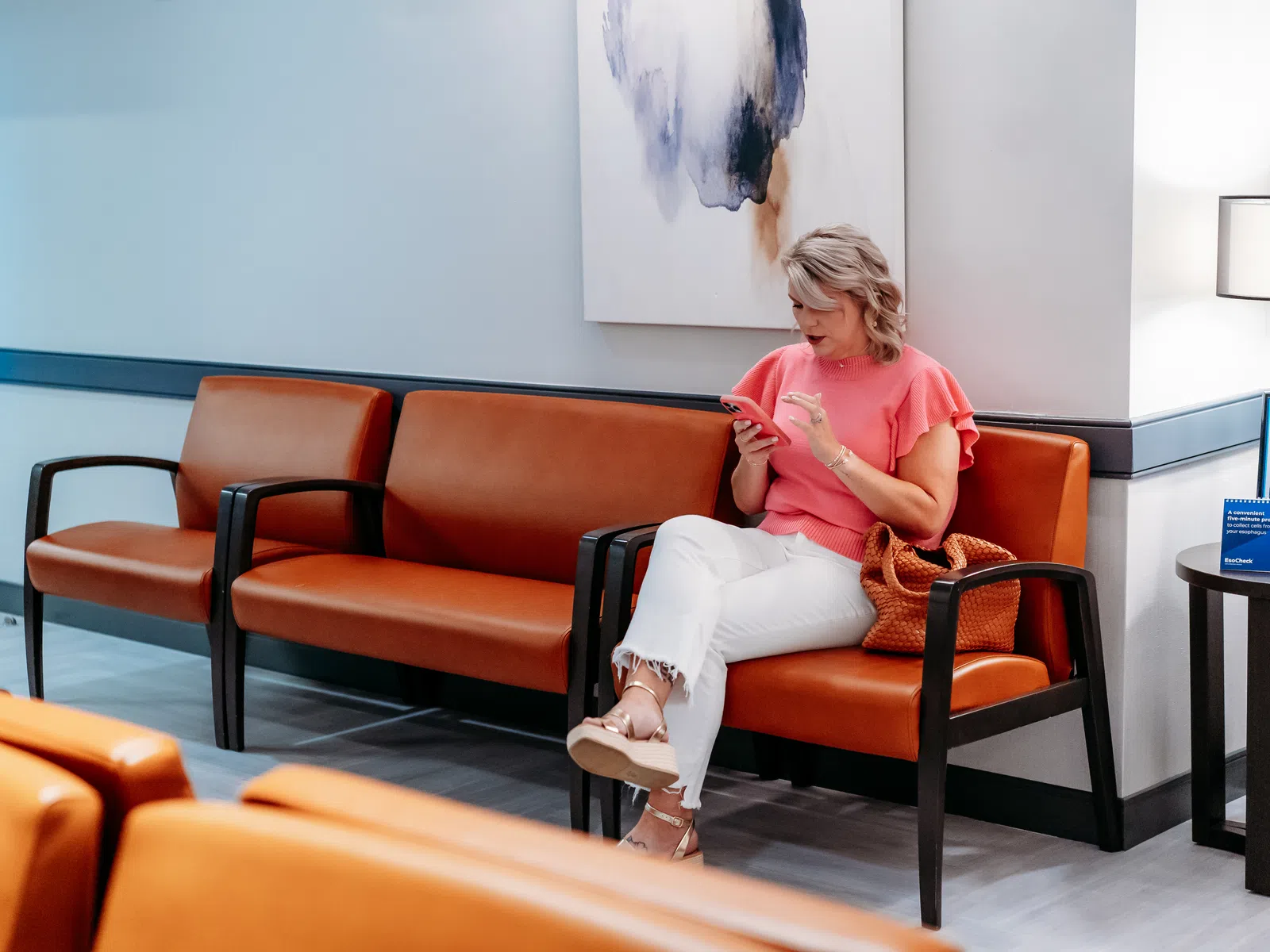 Woman wearing pink top and white pants sitting on brown leather bench in modern waiting room using smartphone