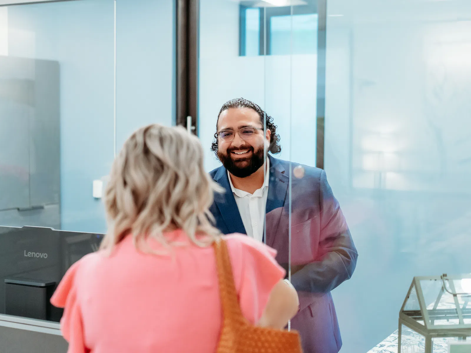Smiling man in office attire speaking through a clear glass partition to a woman in a pink blouse.