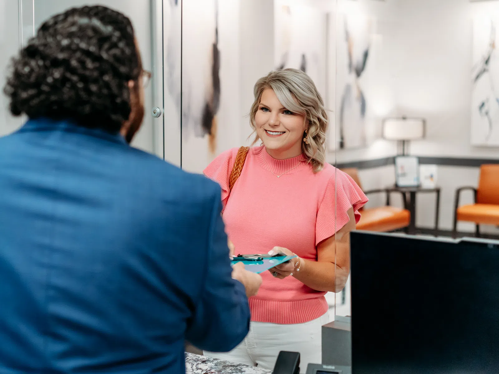 Smiling woman in pink top handing a clipboard to a man in a blue suit at a modern office reception desk.