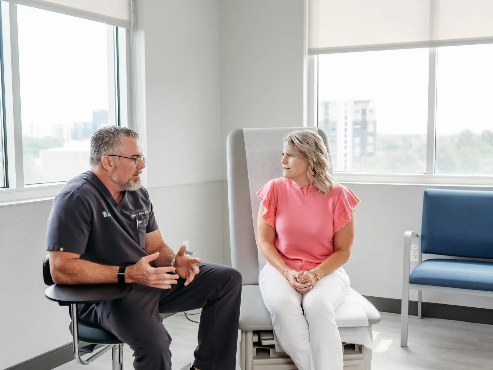 Doctor in black scrubs consulting female patient in pink top in a bright medical office with large windows.